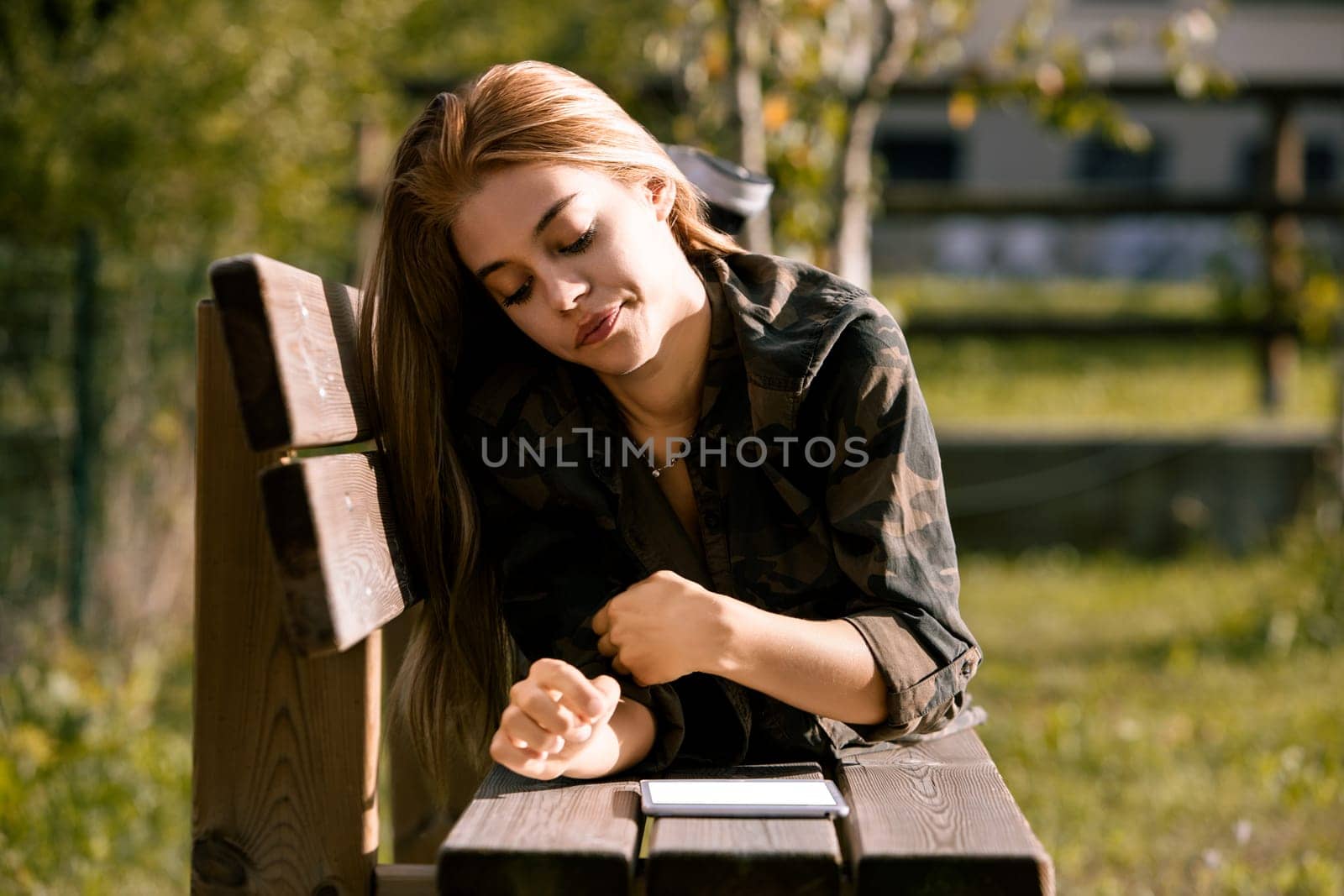 Young woman is lying on her stomach on a wooden bench outside, using a digital tablet. She is wearing a camouflage patterned shirt