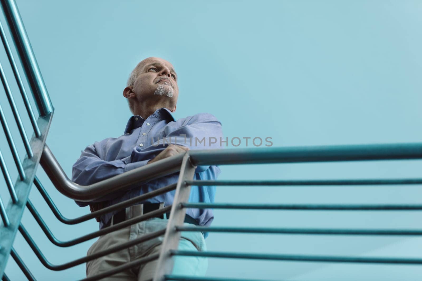 Mature businessman confidently stands on a stairway, arms crossed, looking optimistically towards the future in a blue office setting, embodying leadership, experience, and wisdom