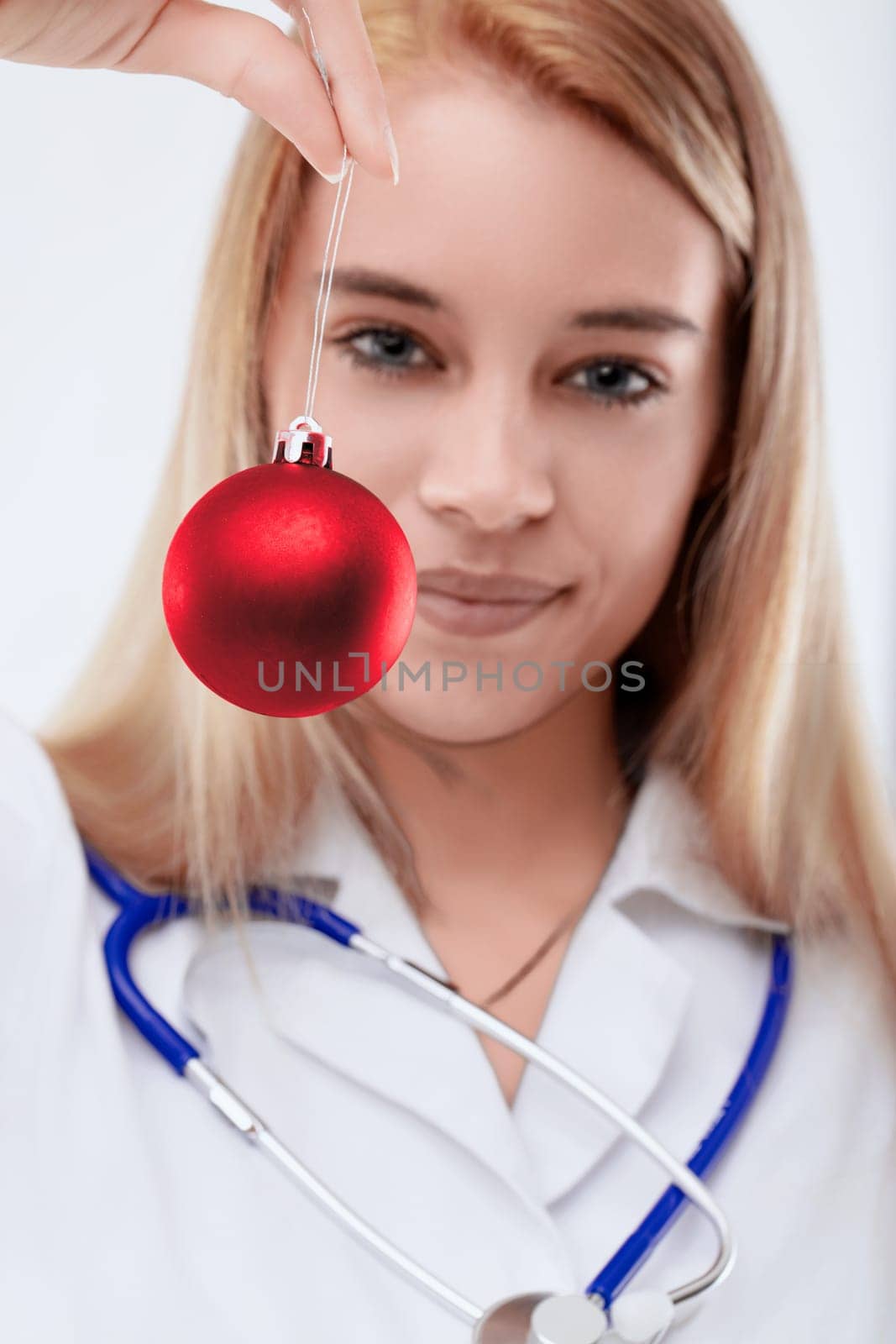 Joyful female doctor smiles holding a red christmas bauble, radiating happiness and festive cheer. Her blonde hair and white background highlight her professionalism and dedication to healthcare