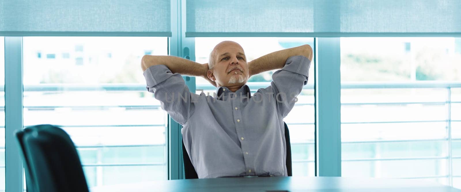 Bald man with a white goatee, dressed in a light blue shirt, reclines in his chair with hands behind his head. The bright, linear background enhances the relaxed scene