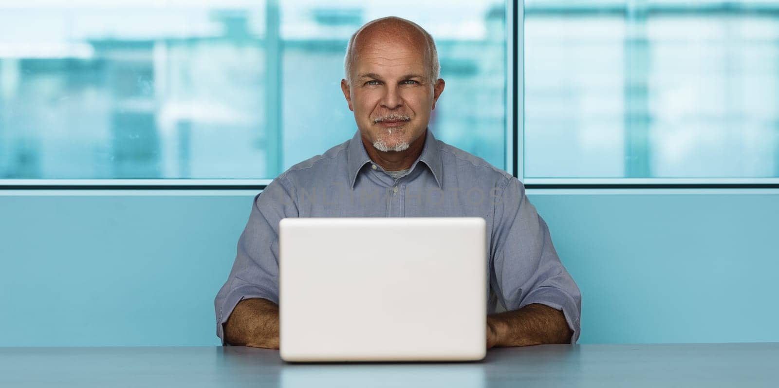 Mature businessman works on his laptop in a modern office space, exuding professionalism and success. Surrounded by a clean workplace with a city view, he appears efficient and productive