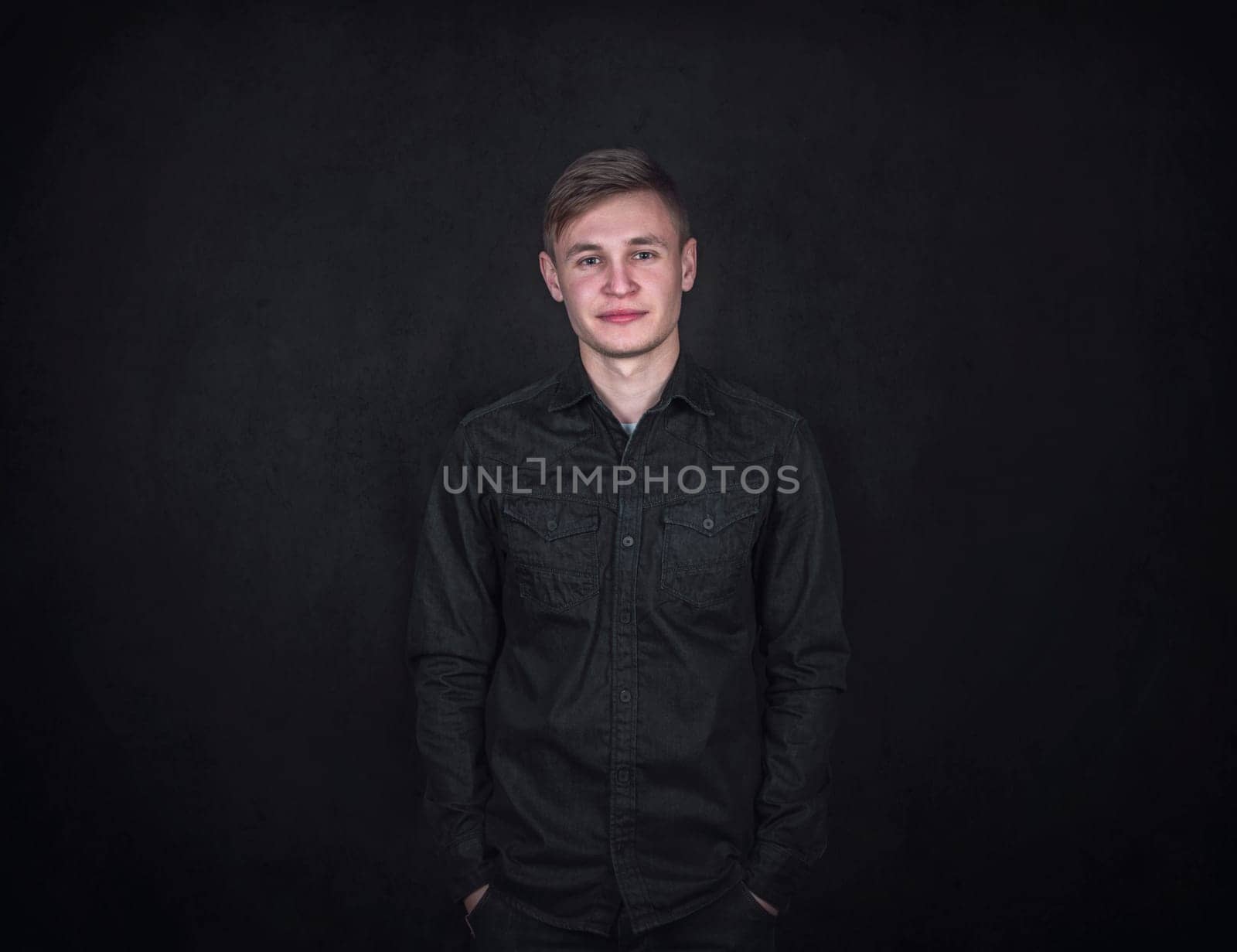 Confident and relaxed young man portrait, keeps hands in pockets against a black wall background. Carefree male student with positive face expression wearing dark blue jeans jacket