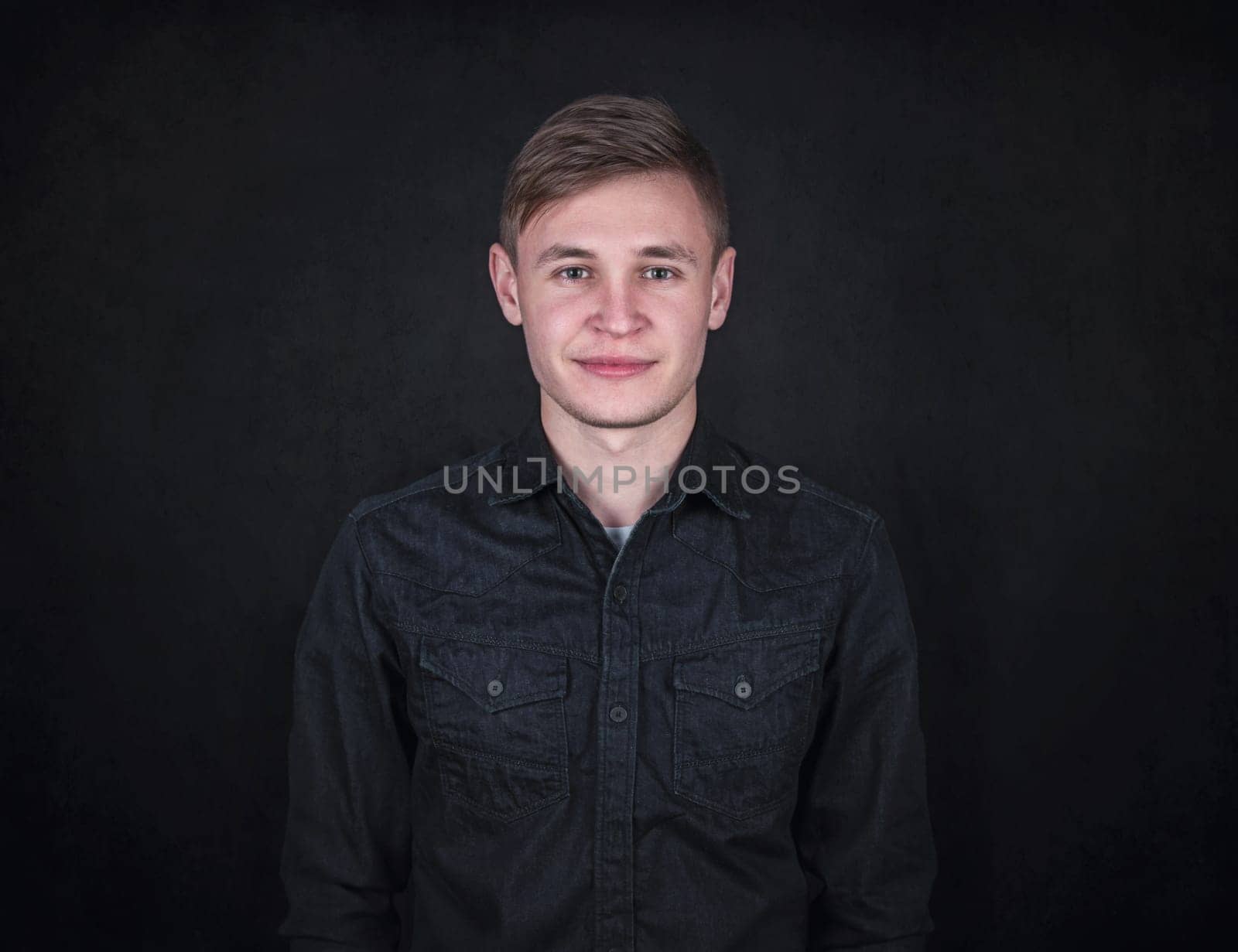 Portrait of a carefree young man against a black wall background looking to camera. Relaxed male model with joyful face expression wearing dark blue jeans jacket