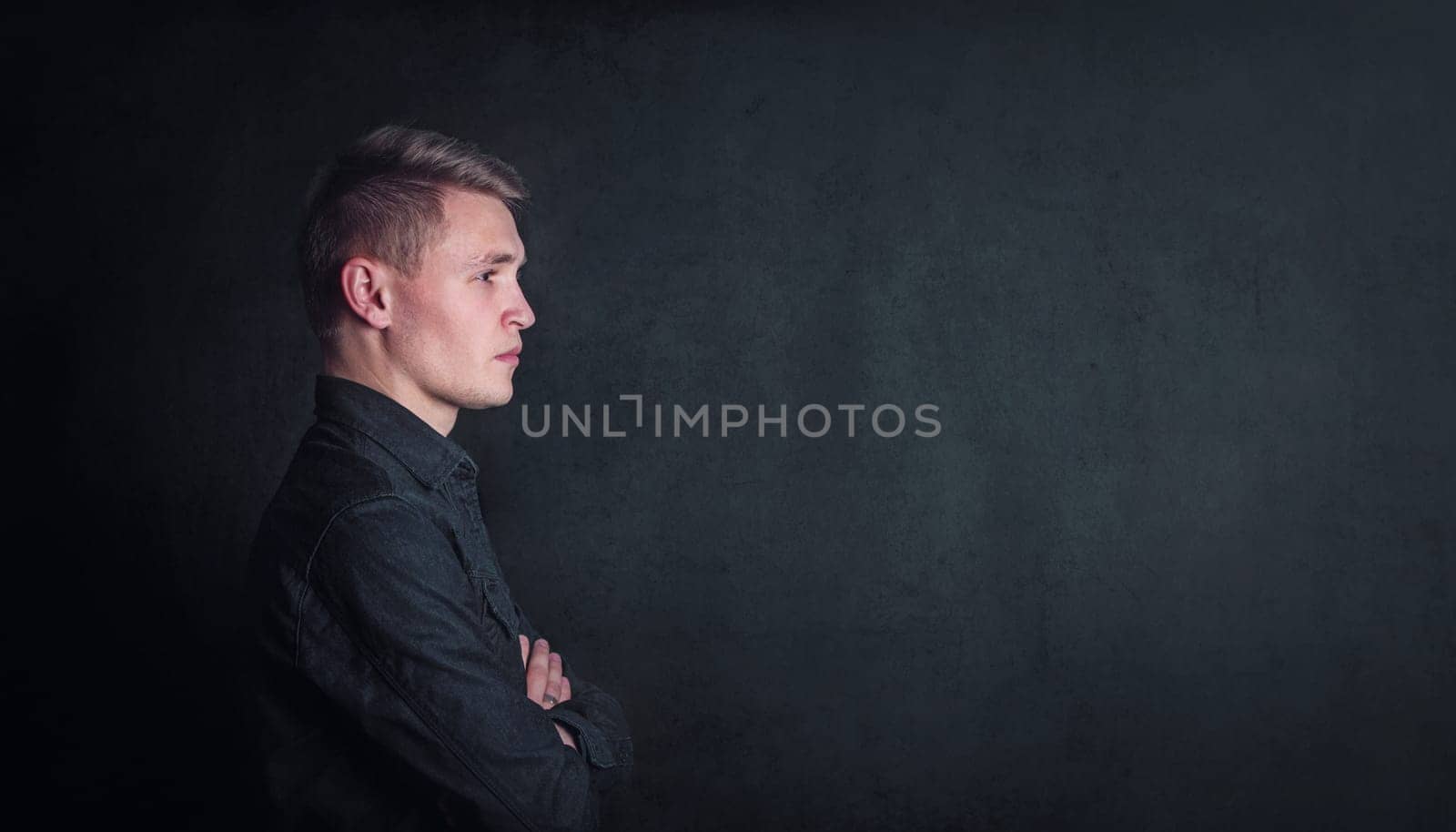Pensive young man side view portrait, keeps arms folded against a dark wall background with copy space. Thoughtful male student with determined face expression