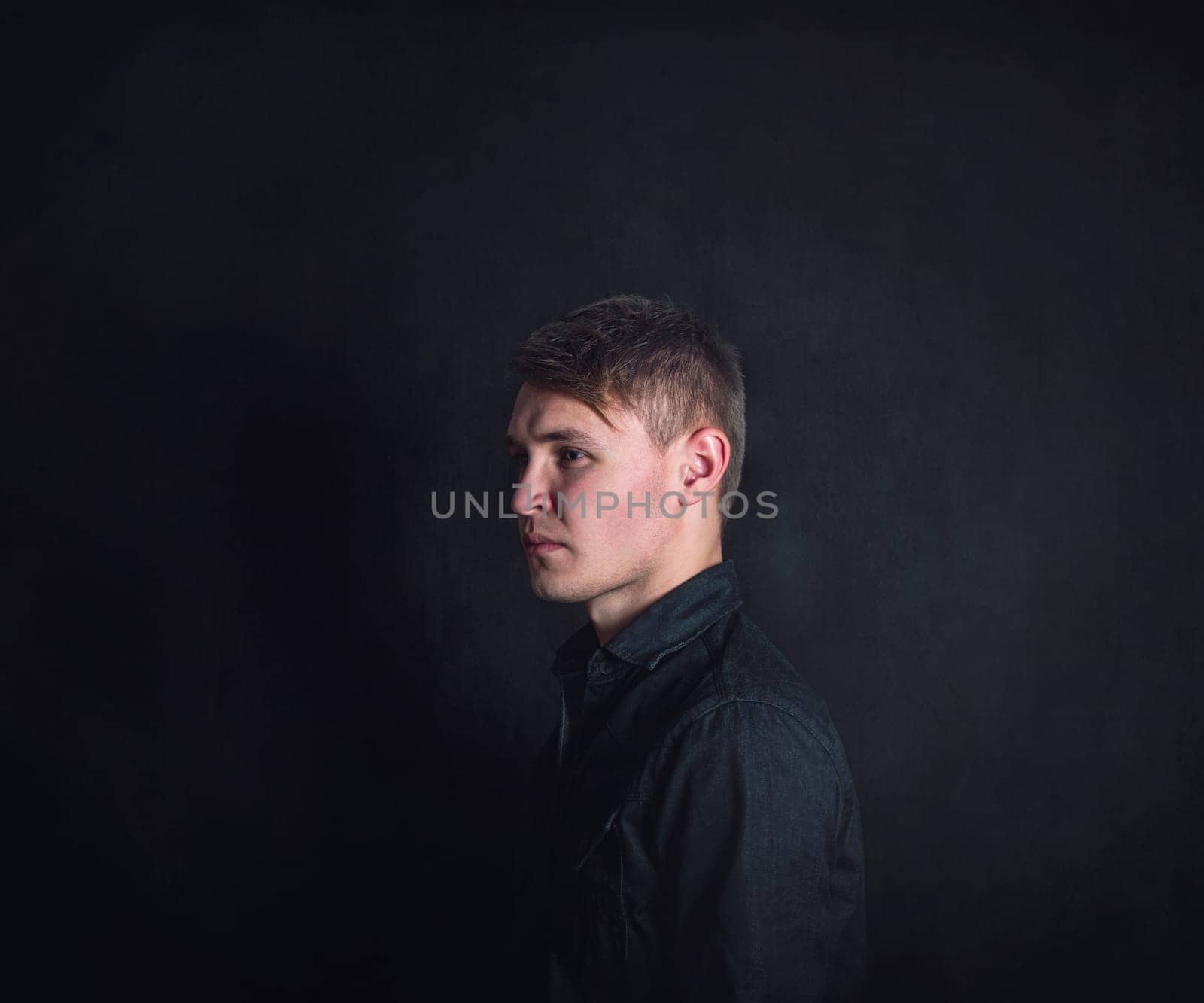 Thoughtful young man side view portrait, looks upset and tired against a dark wall background. Stressed male student feels emotional discomfort and anxiety problems