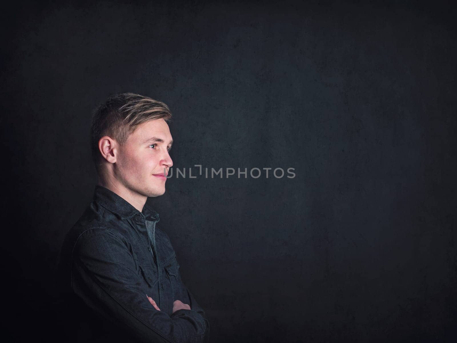 Positive thinking young man side view portrait, looks confident holding arms crossed against a black wall background. Male student reflecting with optimism