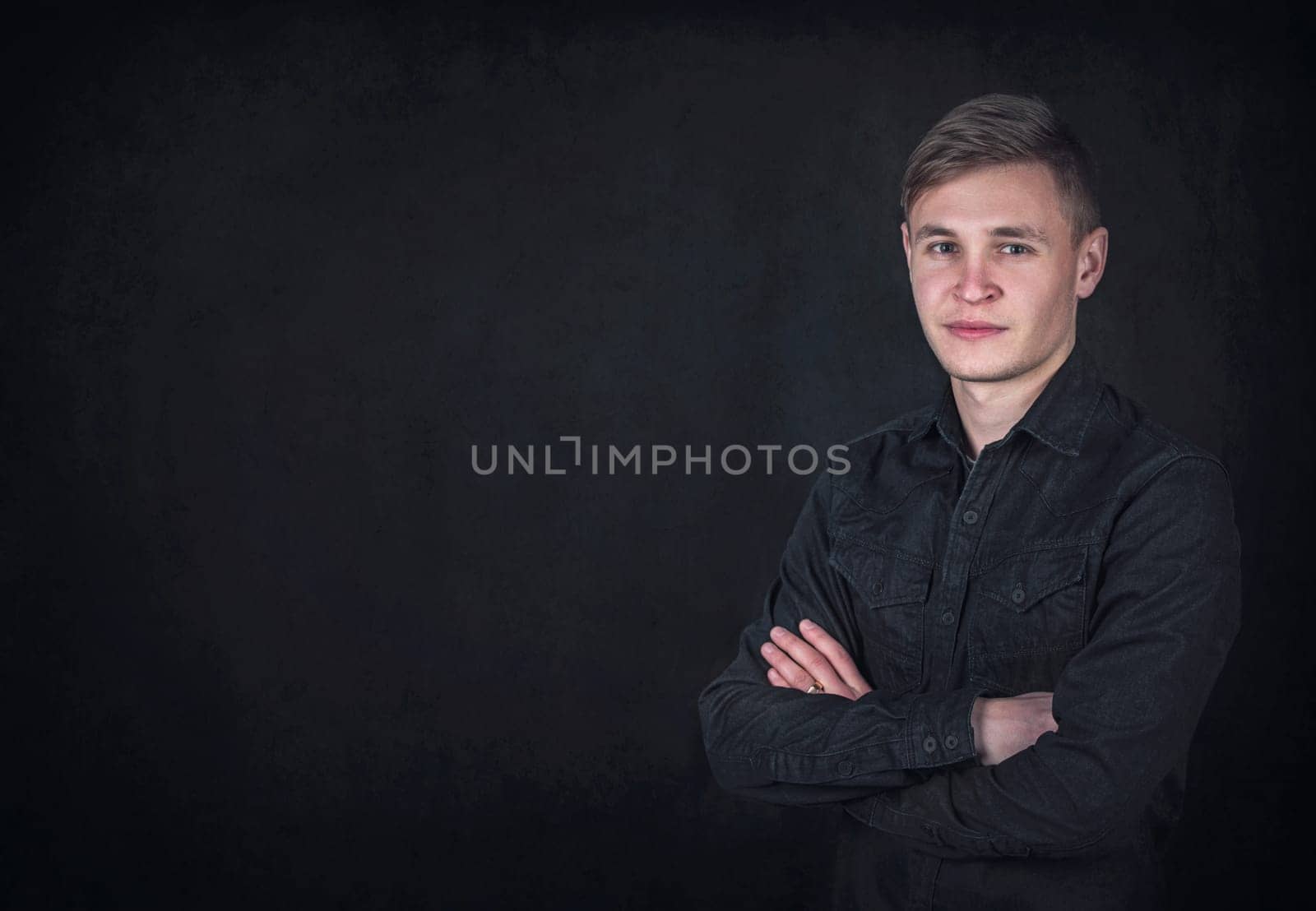 Portrait of a confident young man, keeps arms folded against a black wall background with copy space. Successful male student with determined face expression wearing dark blue jeans jacket
