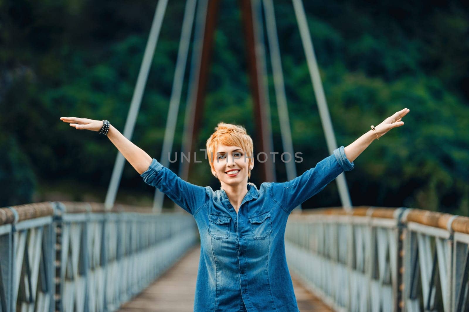 Young woman with short red hair is standing on a bridge with her arms wide open, enjoying the feeling of freedom and happiness. She is wearing a denim shirt and has a big smile on her face