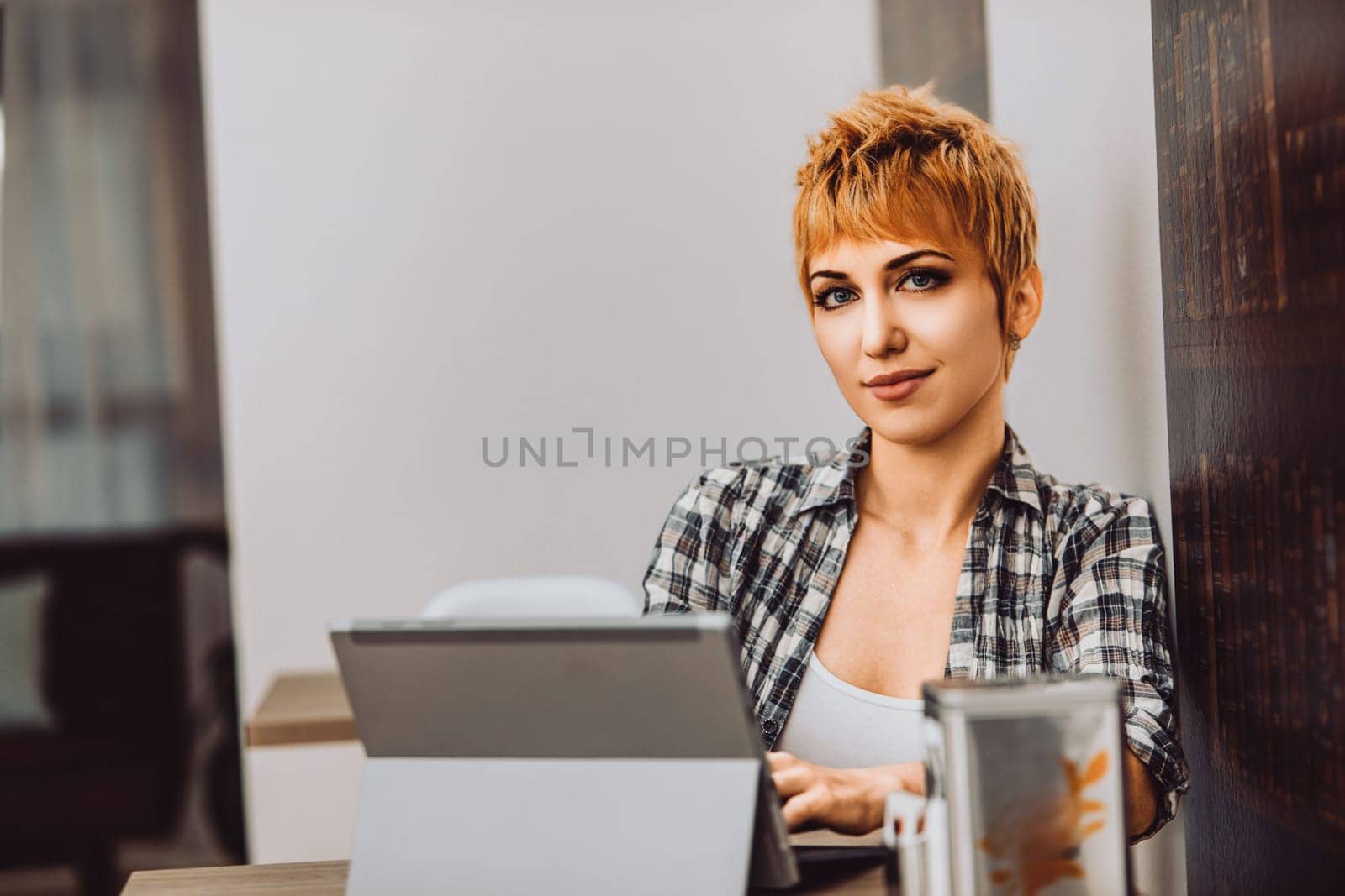 Young woman with short hair is sitting in a cafe, working on her laptop. She is wearing a plaid shirt and smiling slightly