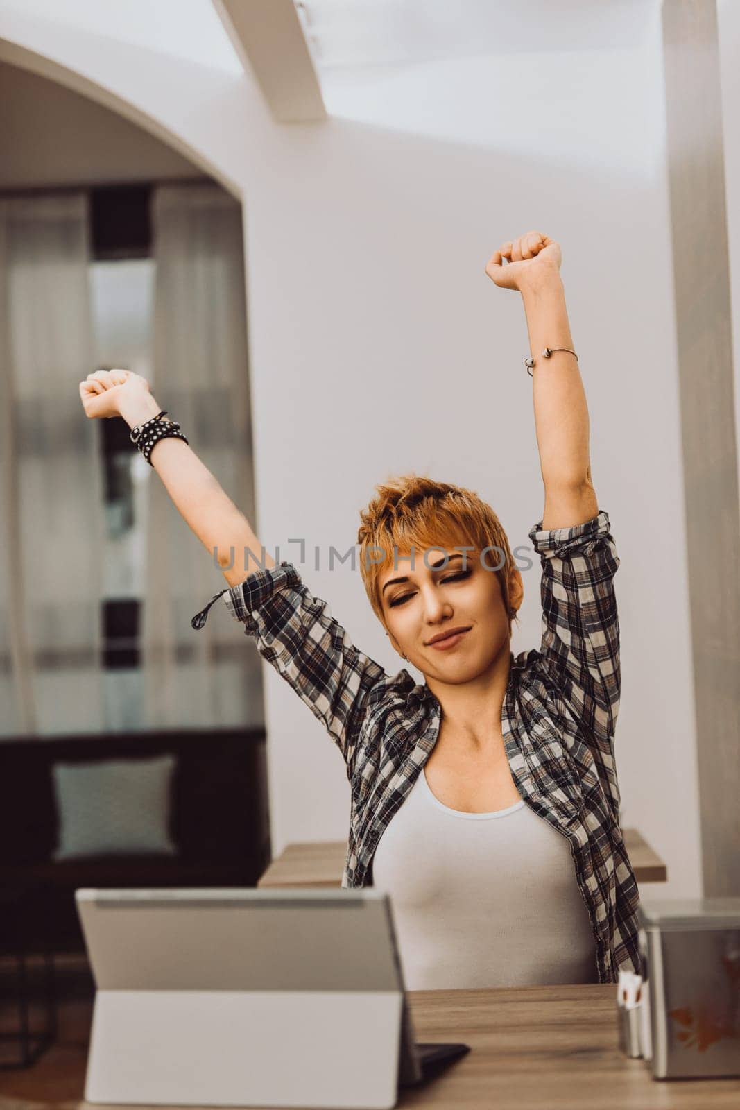 Young woman with short hair and a checkered shirt is stretching her arms while sitting in front of a laptop at a desk