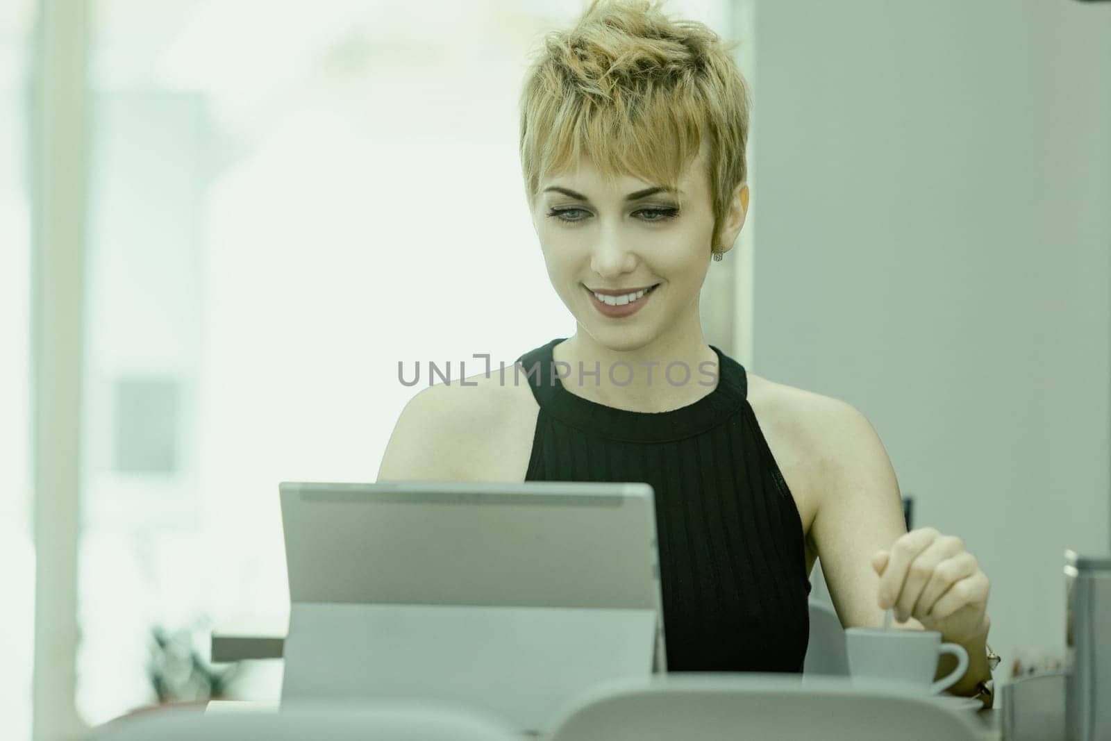 Smiling young woman with short blonde hair works on her laptop in a bright office, exuding confidence and determination