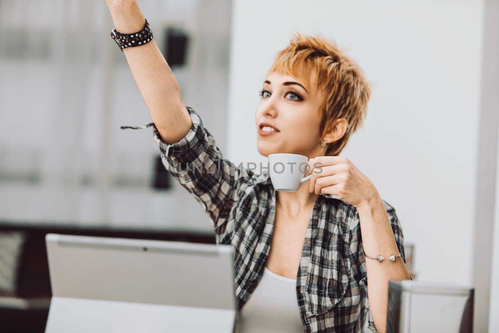 Young casual woman is drinking coffee and pointing with her index finger up while sitting at the table with laptop in a bright room