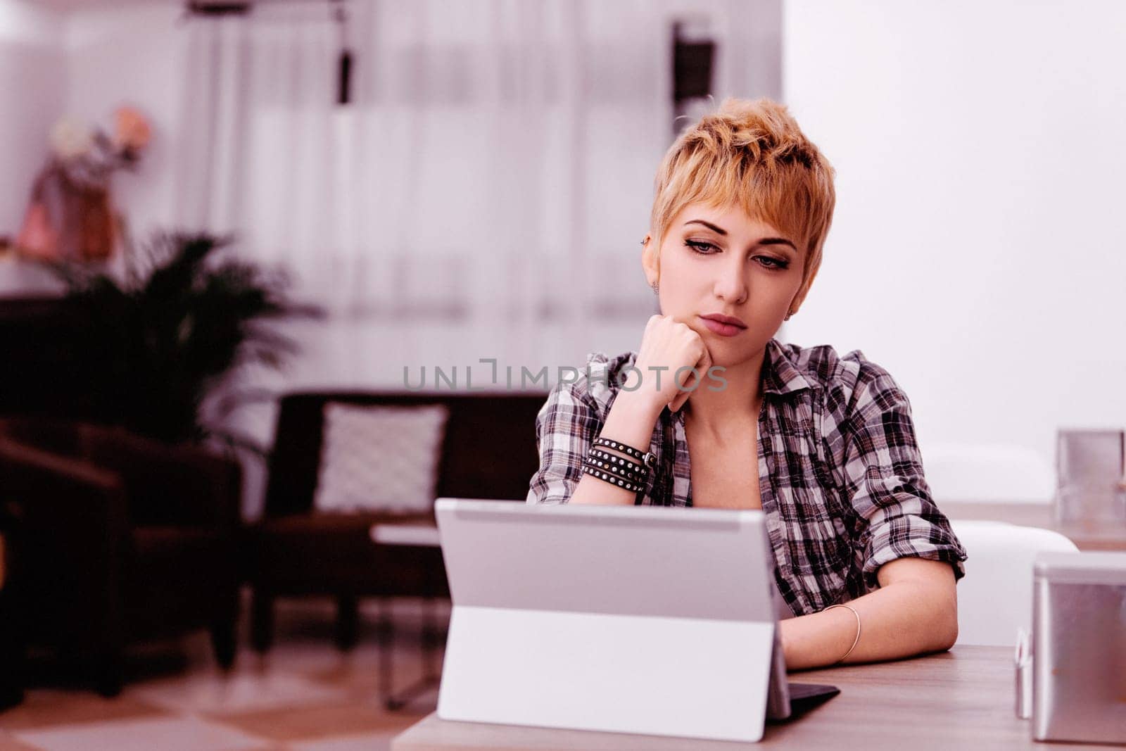 Young woman with short blonde hair looks concerned working from home on tablet, focusing on a problem