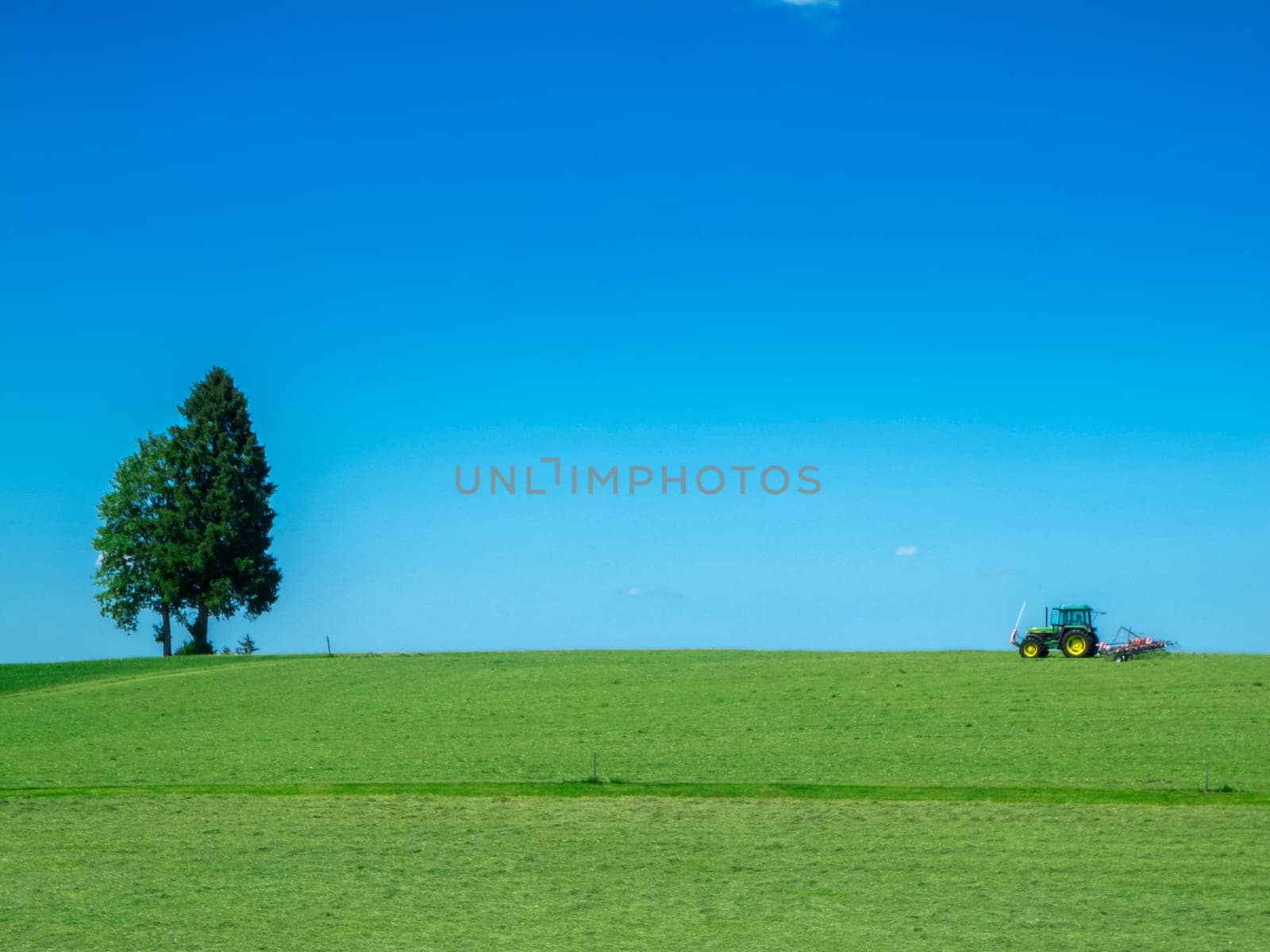 Landscape view of a freshly mown pasture with a single tree and tractor with mowing trailer under a blue sky in Bavaria.