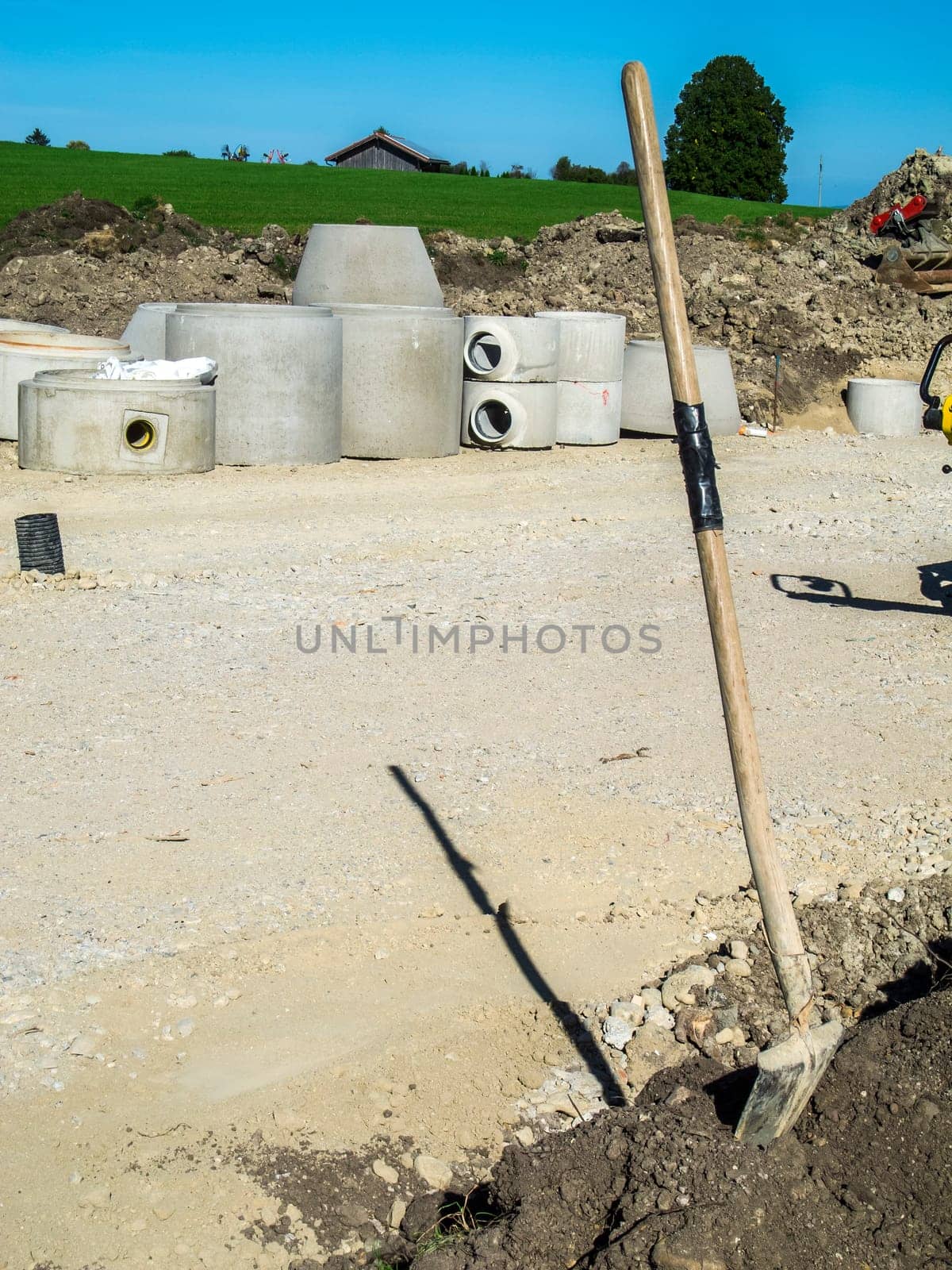 View of a construction site in the Bavarian Allgäu with concrete pipe elements for the construction of sewage canals in the background and a vertical shovel in the foreground in summer sunshine.