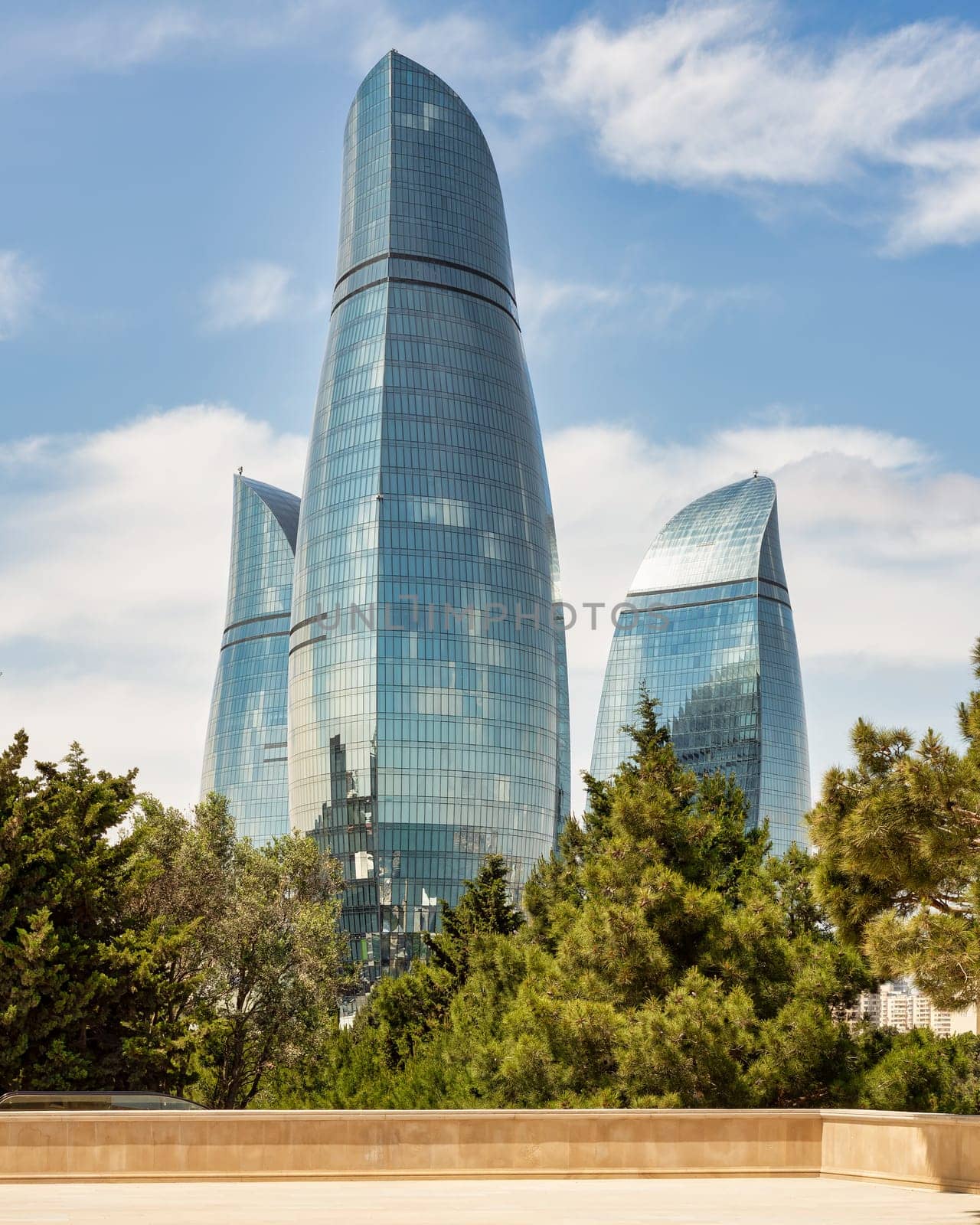 View from the Martyrs Lane of the iconic Flame Towers in Baku, Azerbaijan stand tall against a blue sky, resembling flames. Trees in the foreground add a natural touch to the cityscape