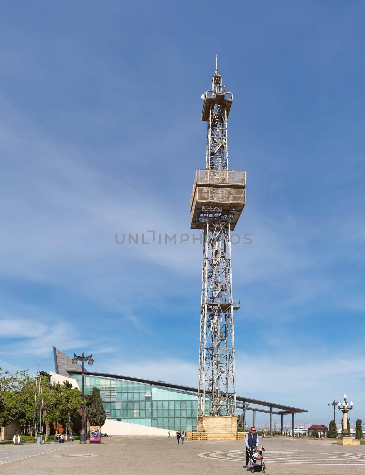 Baku, Azerbaijan - May 10, 2024: Metal parachute tower looms over a paved area with people walking by. Modern business center building is visible in the background