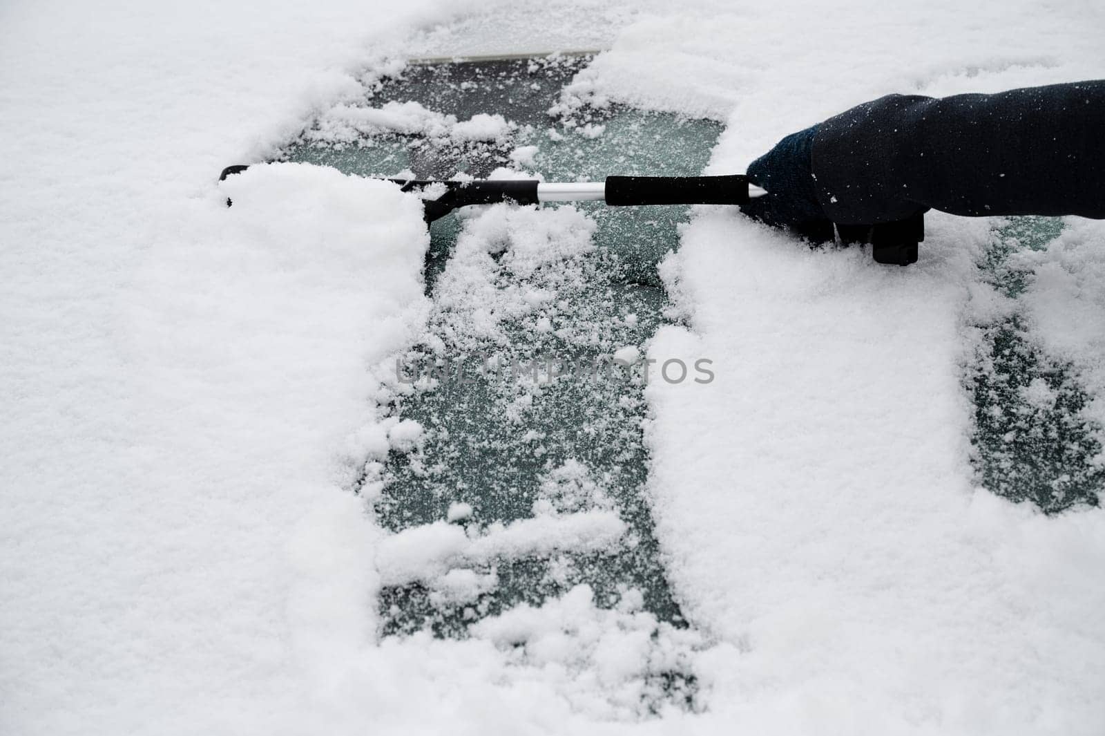 Driver is removing snow from car windshield with ice scraper and broom, clean car window from icy frost, winter season