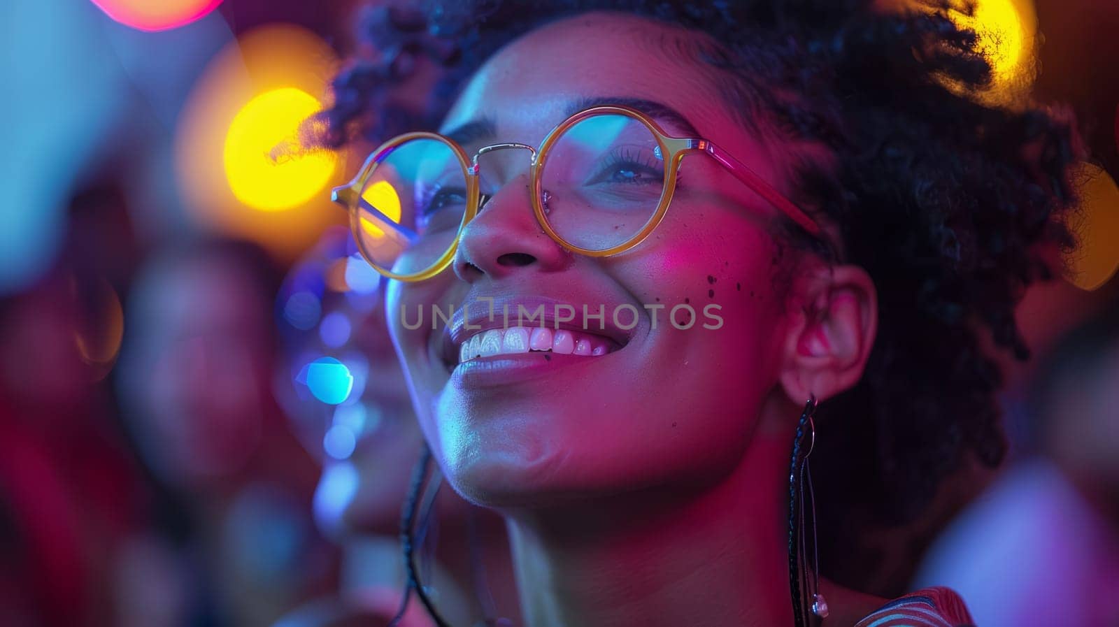 A young woman with glasses smiles brightly, illuminated by colorful lights in a nighttime setting