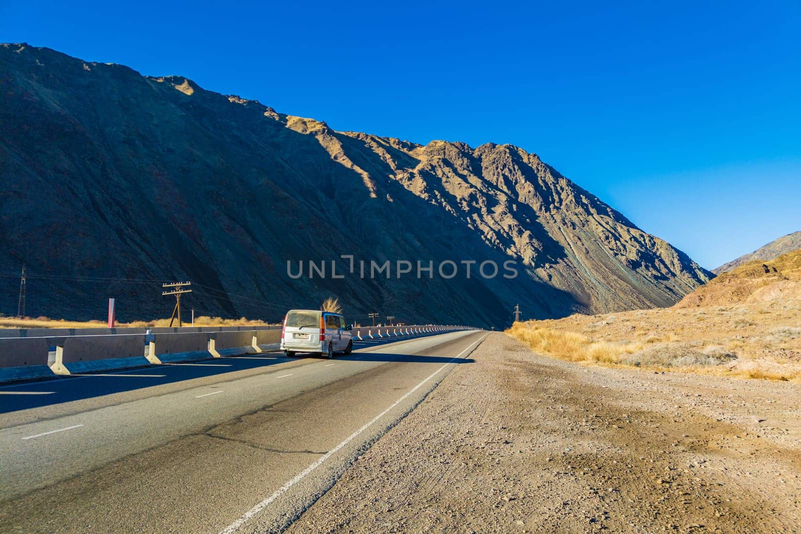 Car is driving along the road through natural mountain landscape with concrete barrier at sunny autumn day in Boom Gorge, Kyrgyzstan - December 16, 2022