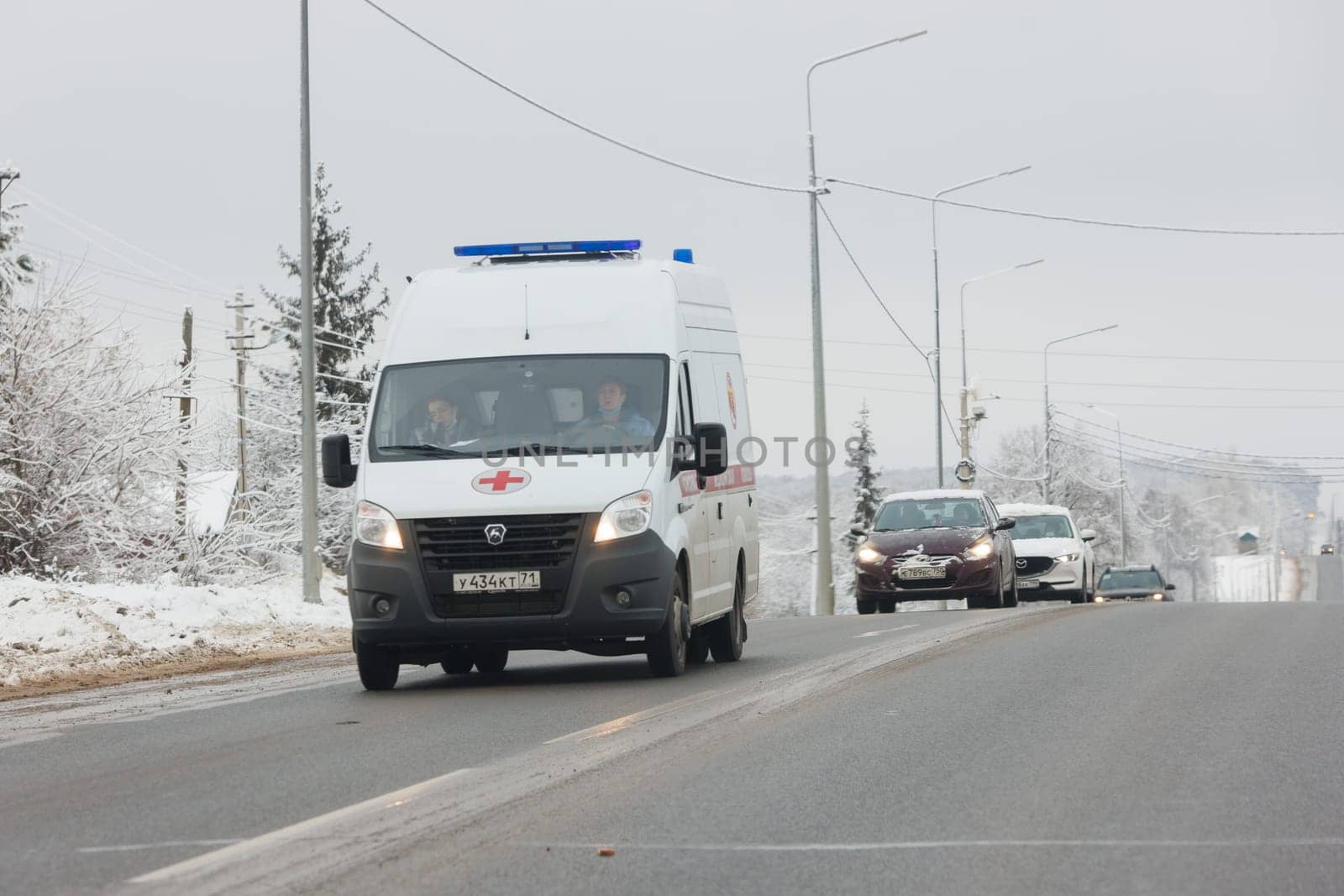 white ambulance minibus with two women inside the cabin driving along winter road at cloudy day in Tula, Russia - December 9. 2021