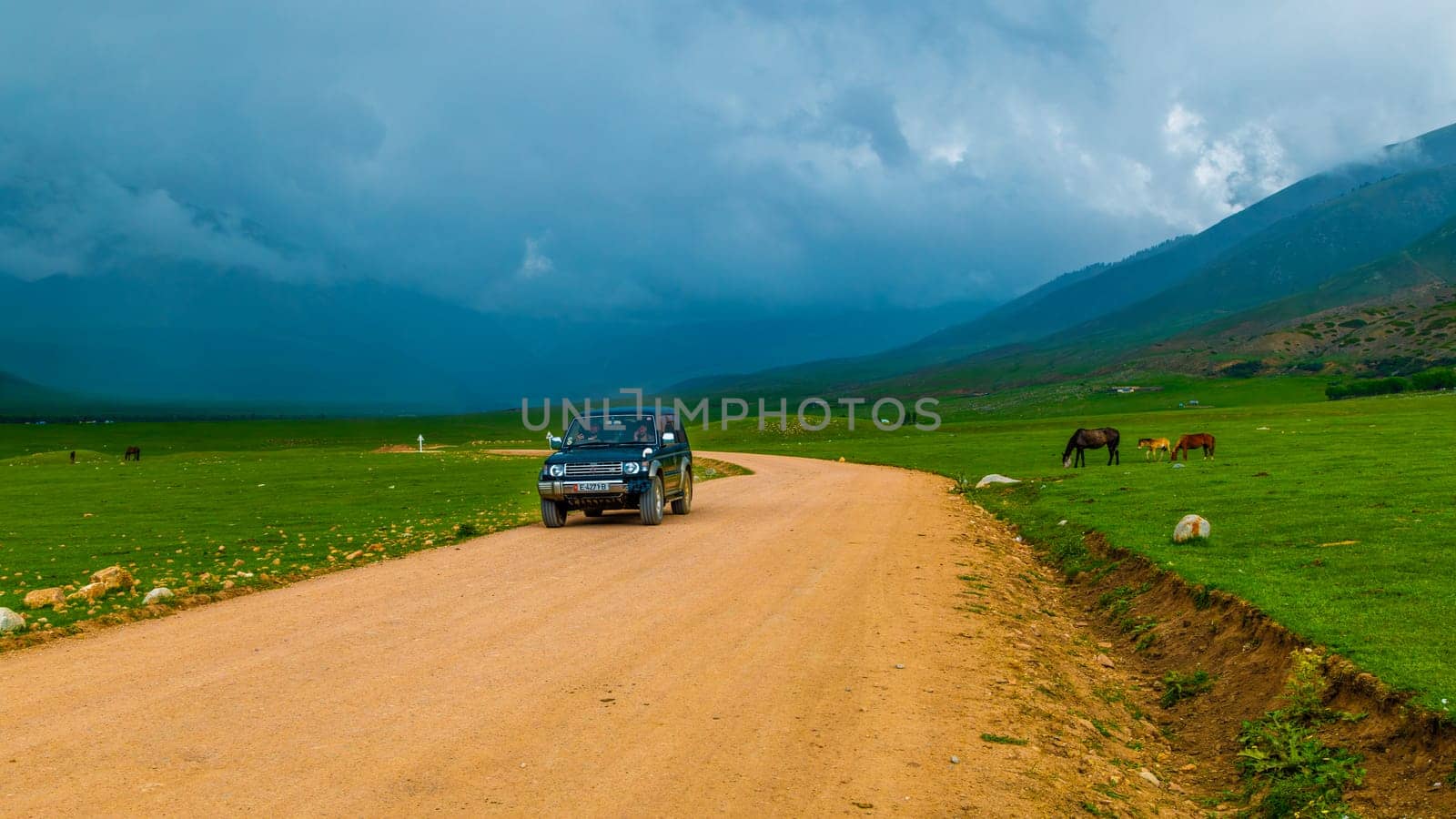 old SUV driving along dry dirt road near grazing horses before thunderstorm in Semenovskoye gorge, Kyrgyzstan at July 5, 2023