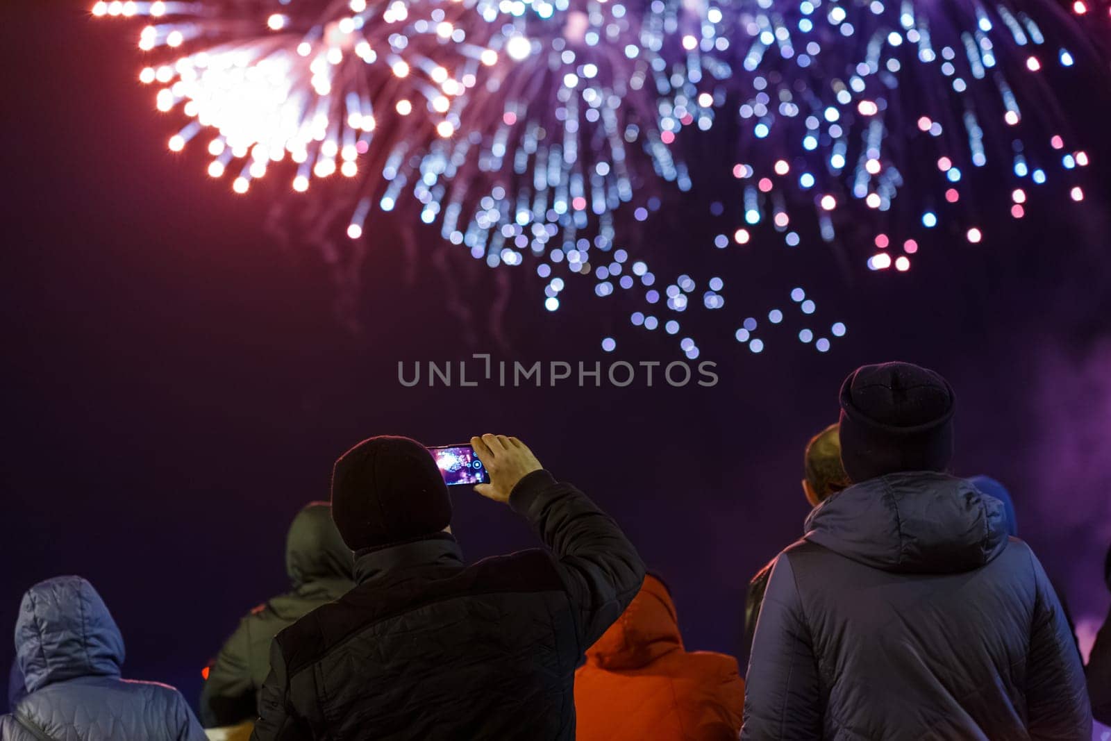 group of unrecognizable people in warm clothes are watching fireworks in a night sky, rear view