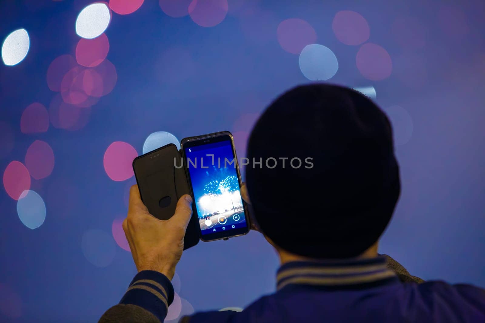 unrecognizable man with black hat photographing fireworks at night with cellphone, closeup with selective focus, rear view