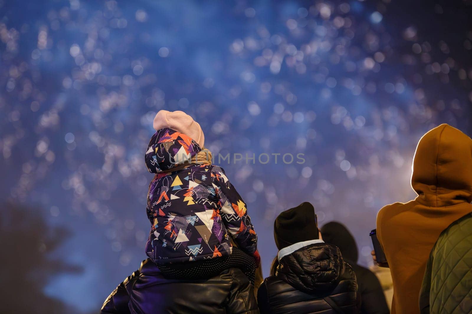group of unrecognizable people with a child on shoulders in warm clothes are watching fireworks in a night sky, rear view