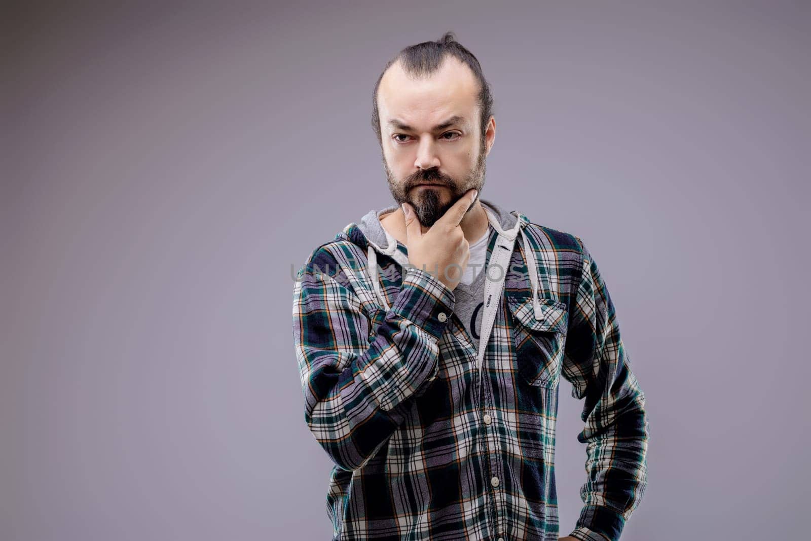 Bearded man wearing a plaid shirt is holding his chin with a thoughtful expression, trying to make a decision or find a solution
