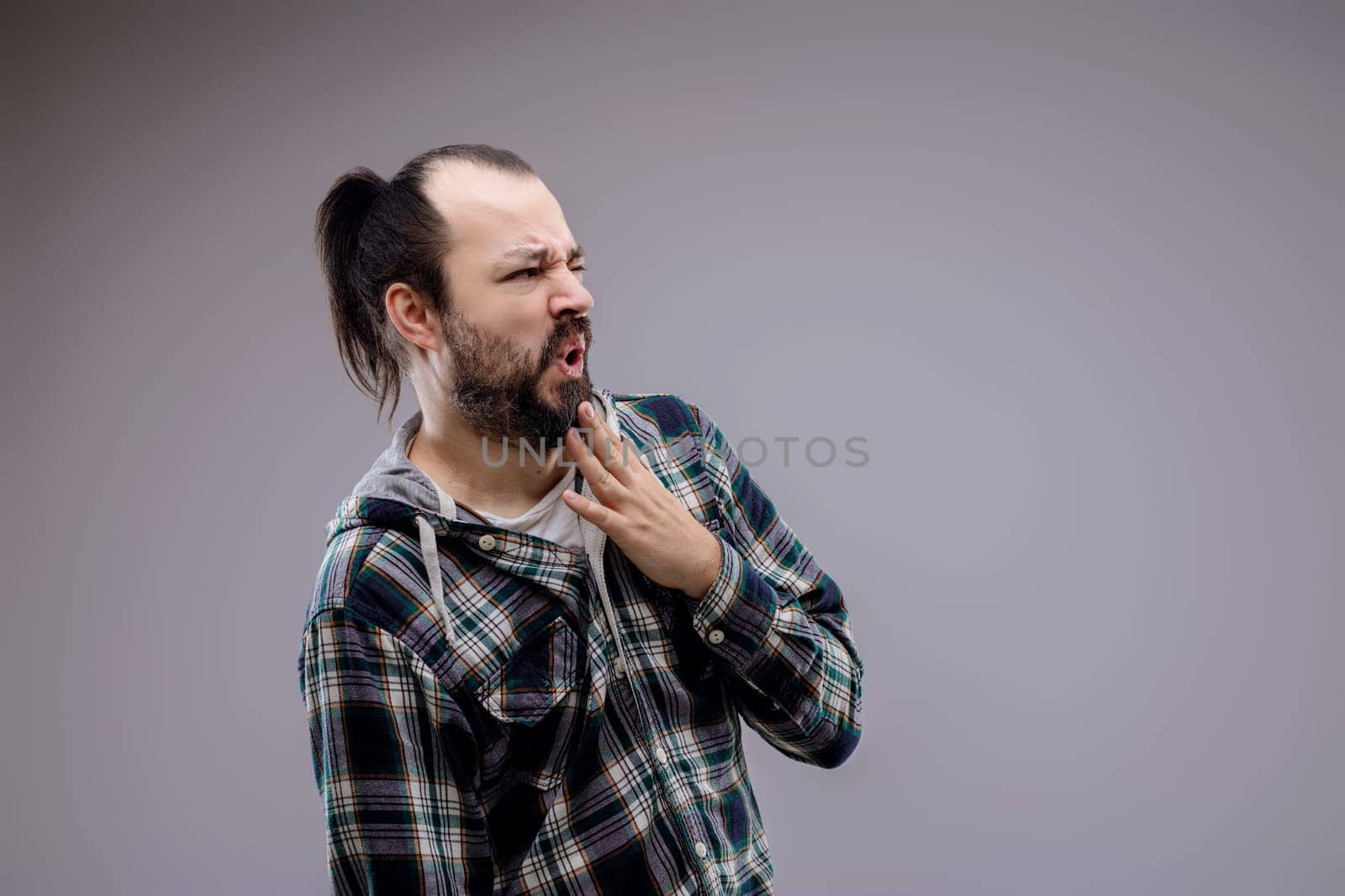 Casual man with beard and ponytail wearing a plaid shirt, touching his face with a disgusted expression on a neutral background