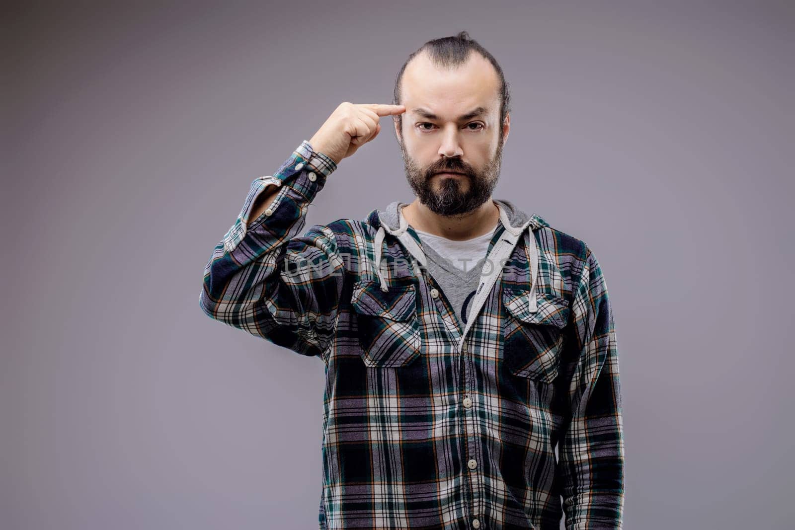 Bearded man in a checkered shirt looks thoughtful, pointing to his temple with a serious expression, showing a mix of emotions like doubt and stress