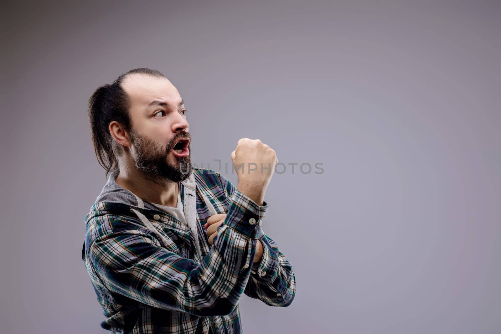 Portrait of a man with a ponytail and beard, wearing a plaid shirt, looking furious while shouting and gesturing with his fists