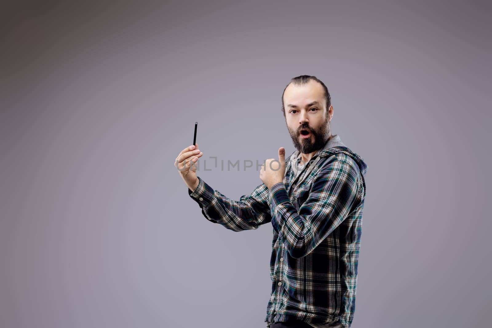 Surprised man takes a selfie with a smartphone, showing happiness and wonder. He gives a thumbs up in a studio with a gray background, wearing a casual checkered shirt