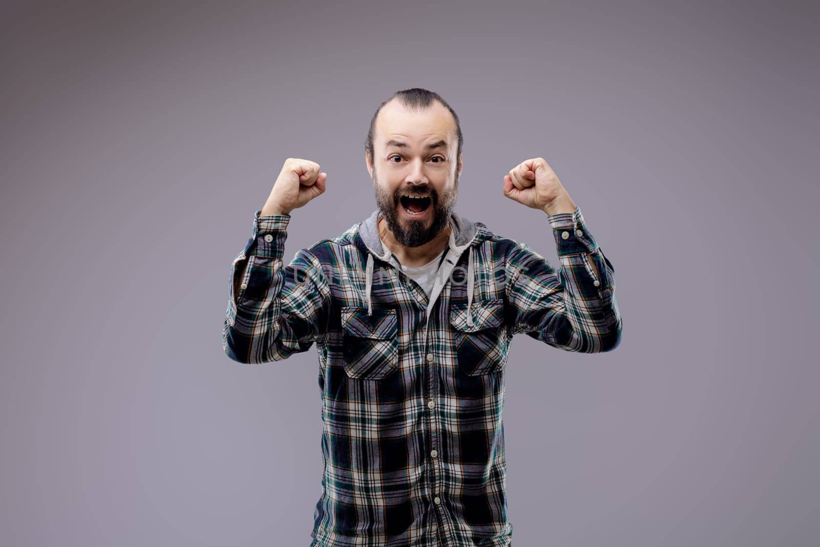 Man with a beard is expressing positive feelings by raising his fists in the air and opening his mouth wide while standing in front of a grey background