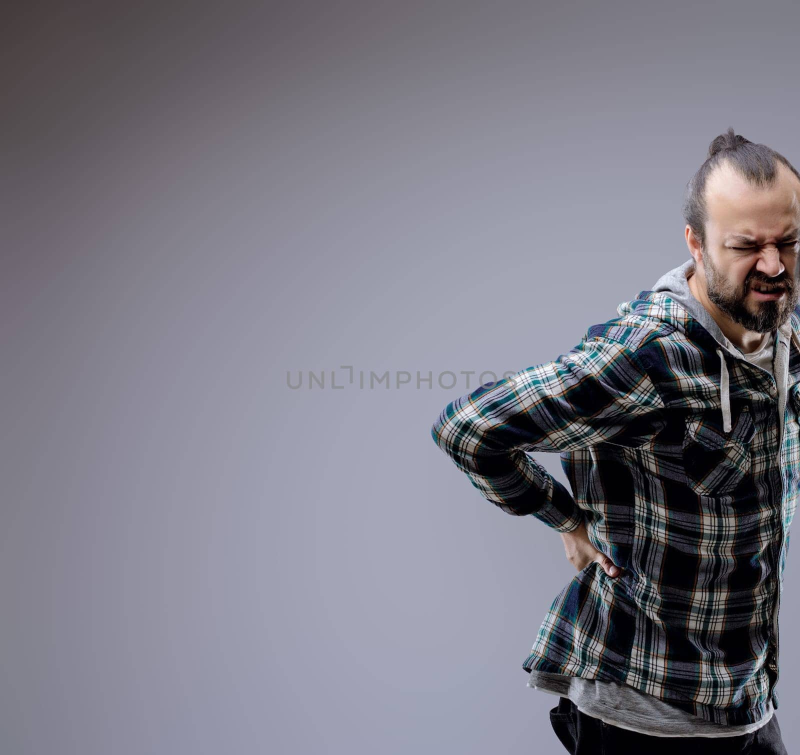 Man in his 30s grimacing in pain, holding his lower back due to backache, showcasing negative emotions in a studio shot against a gray background