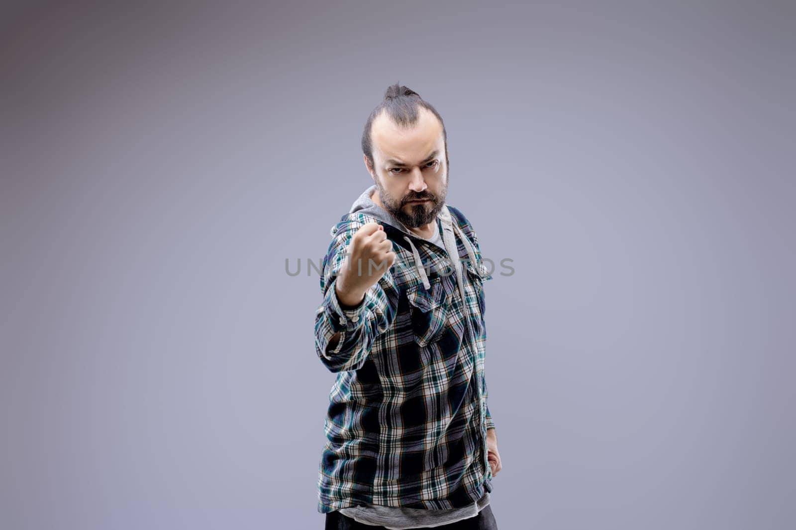 Aggressive man with a beard wearing a plaid shirt is throwing a punch with a threatening facial expression on a gray background