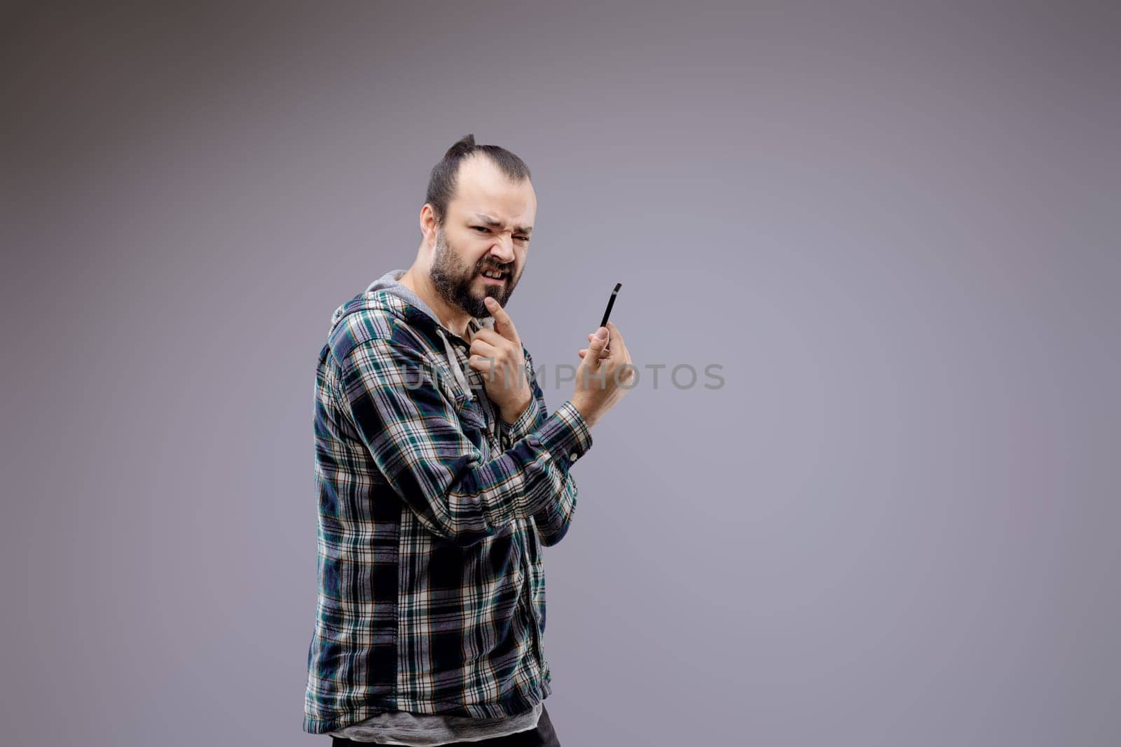 Man with a beard is holding a pen and looking suspicious while making a face. He is standing in front of a grey background