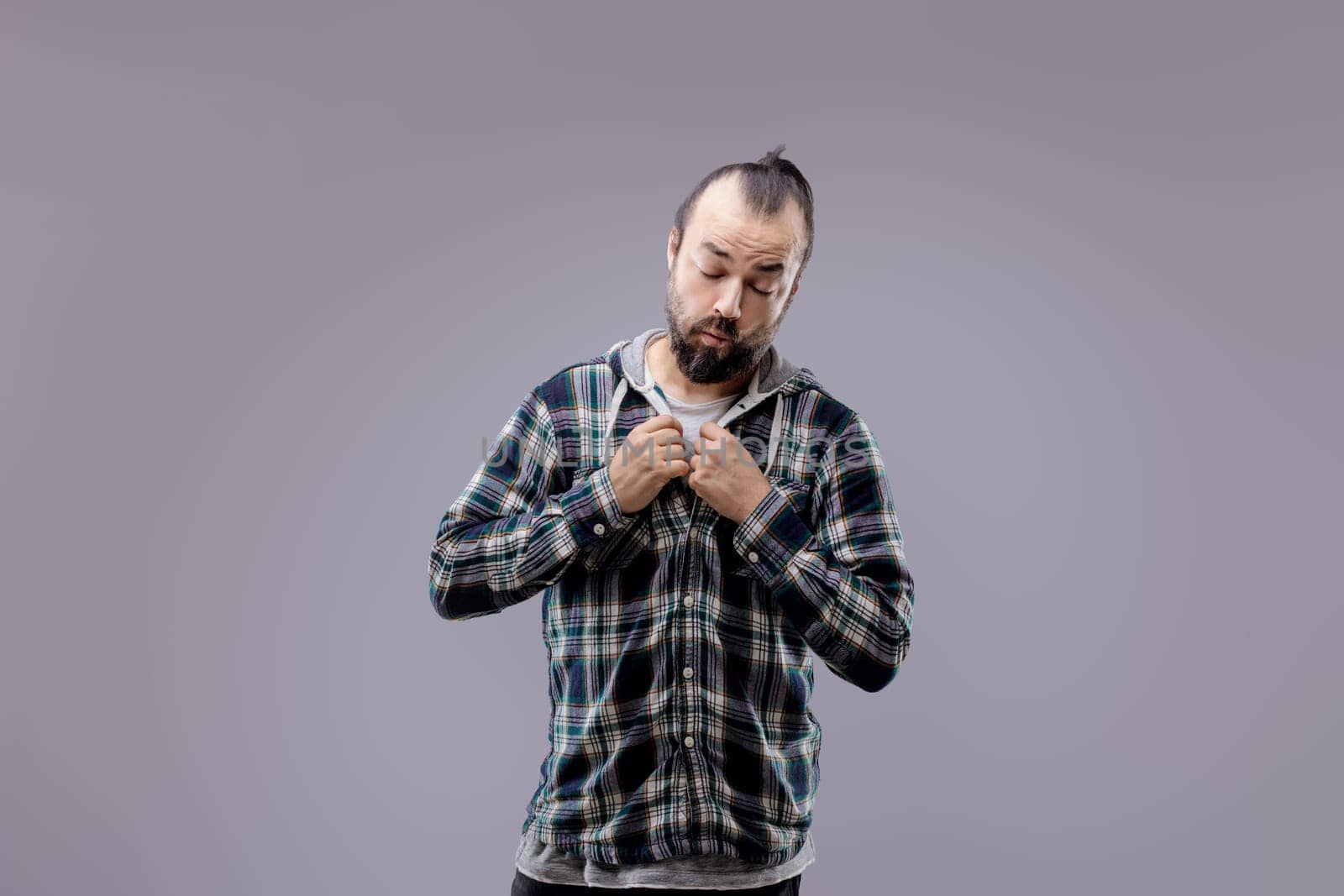 Studio portrait of a bearded man with his hair tied back, wearing a plaid shirt over a hooded sweatshirt, buttoning his shirt with a thoughtful expression