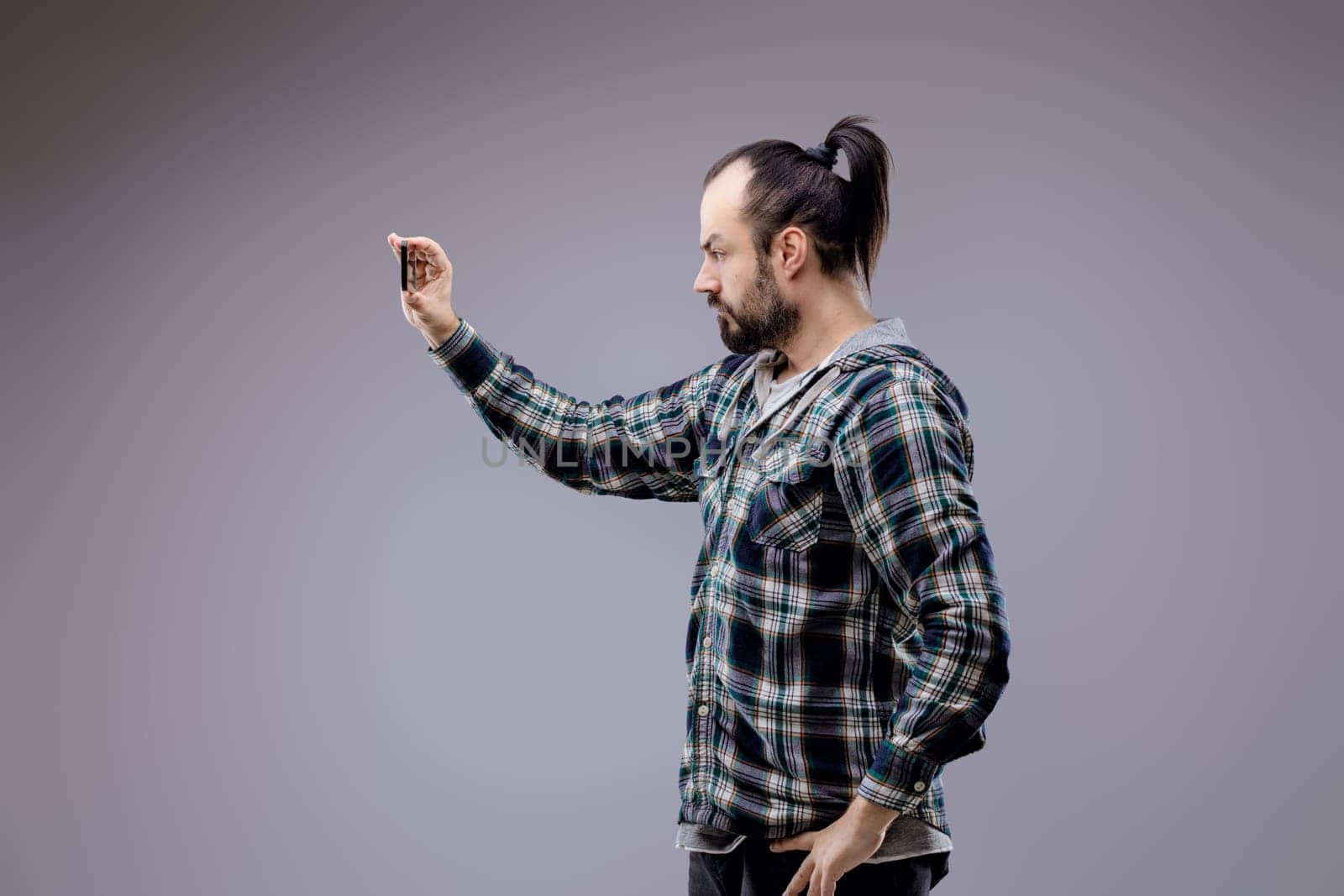 Casually dressed man takes a selfie with a smartphone against a gray background, showcasing modern lifestyle and technology