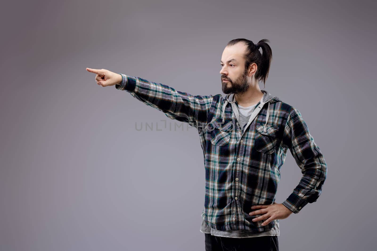 Bearded man pointing accusingly, hand on hip, with a serious expression showing conflict, frustration, and anger, exuding assertiveness and confidence against a gray background
