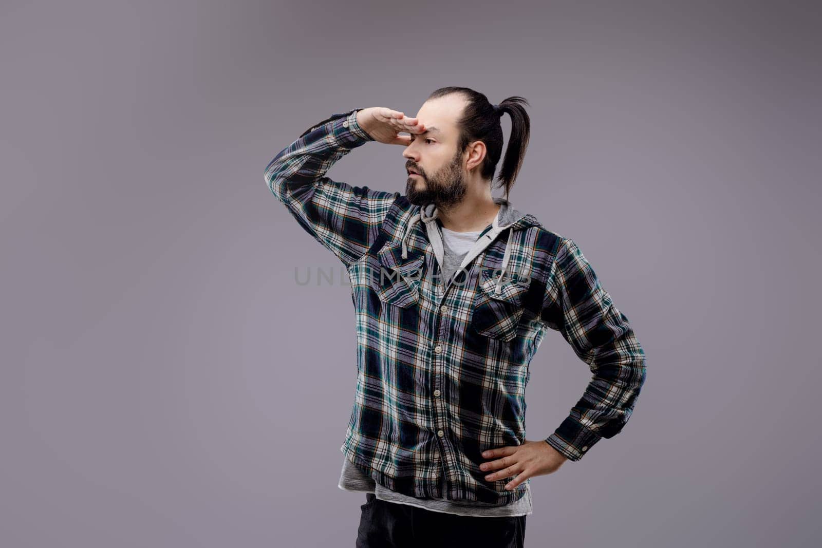 Man with a beard and ponytail is standing with one hand on his hip and the other shielding his eyes as he looks intently into the distance