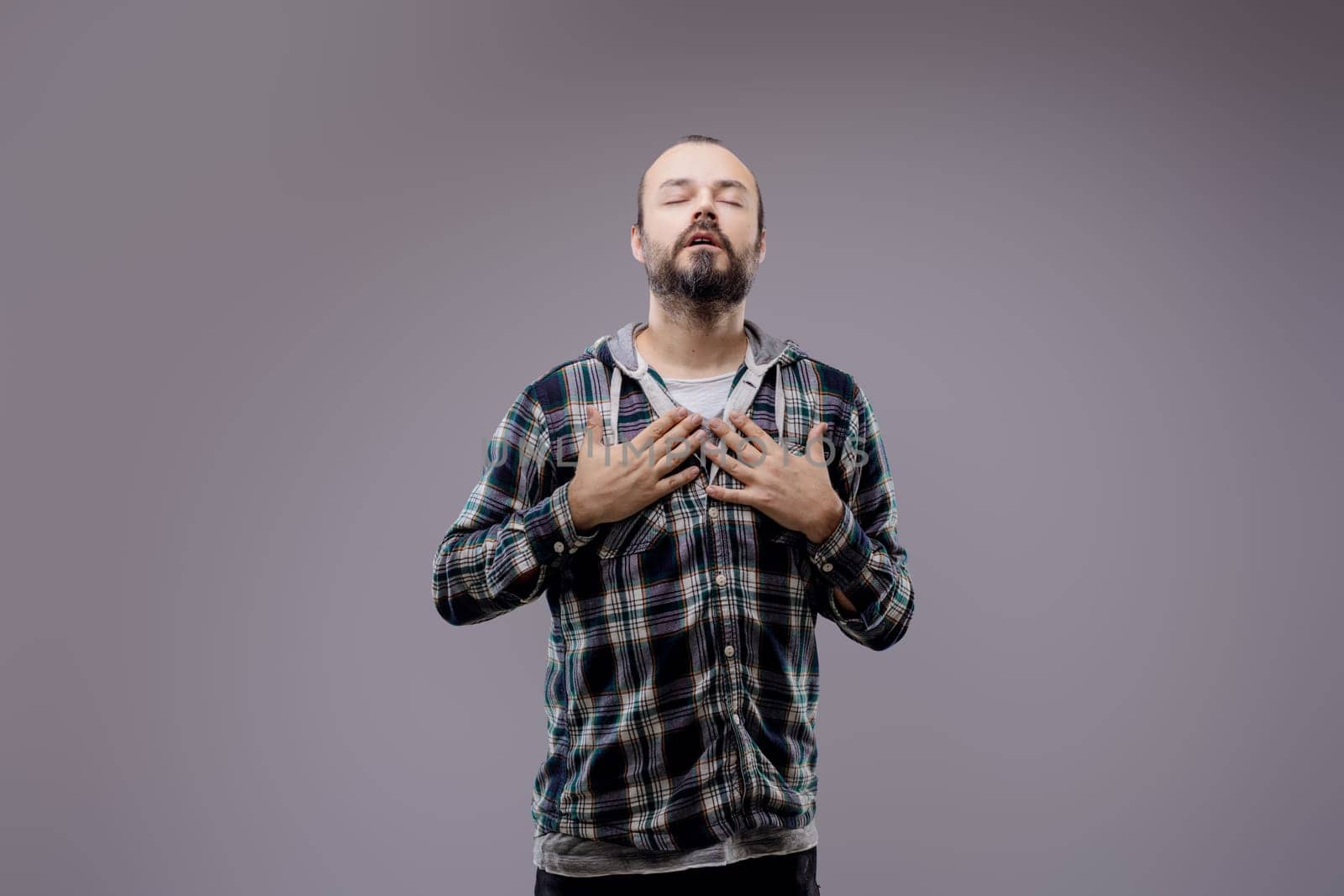 Bearded man with closed eyes holding both hands on his chest, experiencing discomfort and pain. He is standing on a gray background