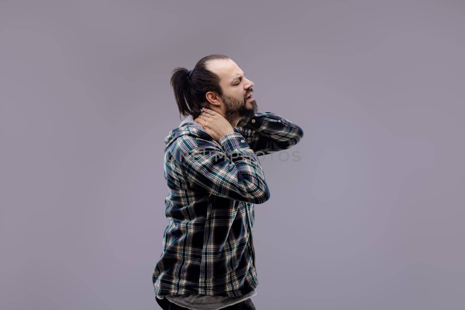 Casual man with beard and ponytail wearing a plaid shirt, standing and suffering from neck pain, massaging his neck with a painful expression