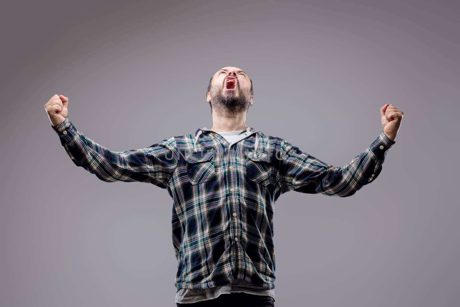 Bearded man celebrates victory with raised fists, showing pure joy and excitement against a gray studio background
