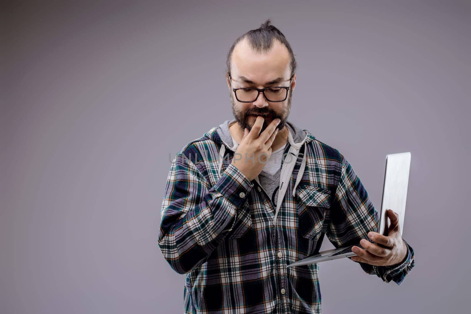Focused web developer with laptop contemplates coding problem in studio. Gray background enhances serious atmosphere. Expertise and determination evident