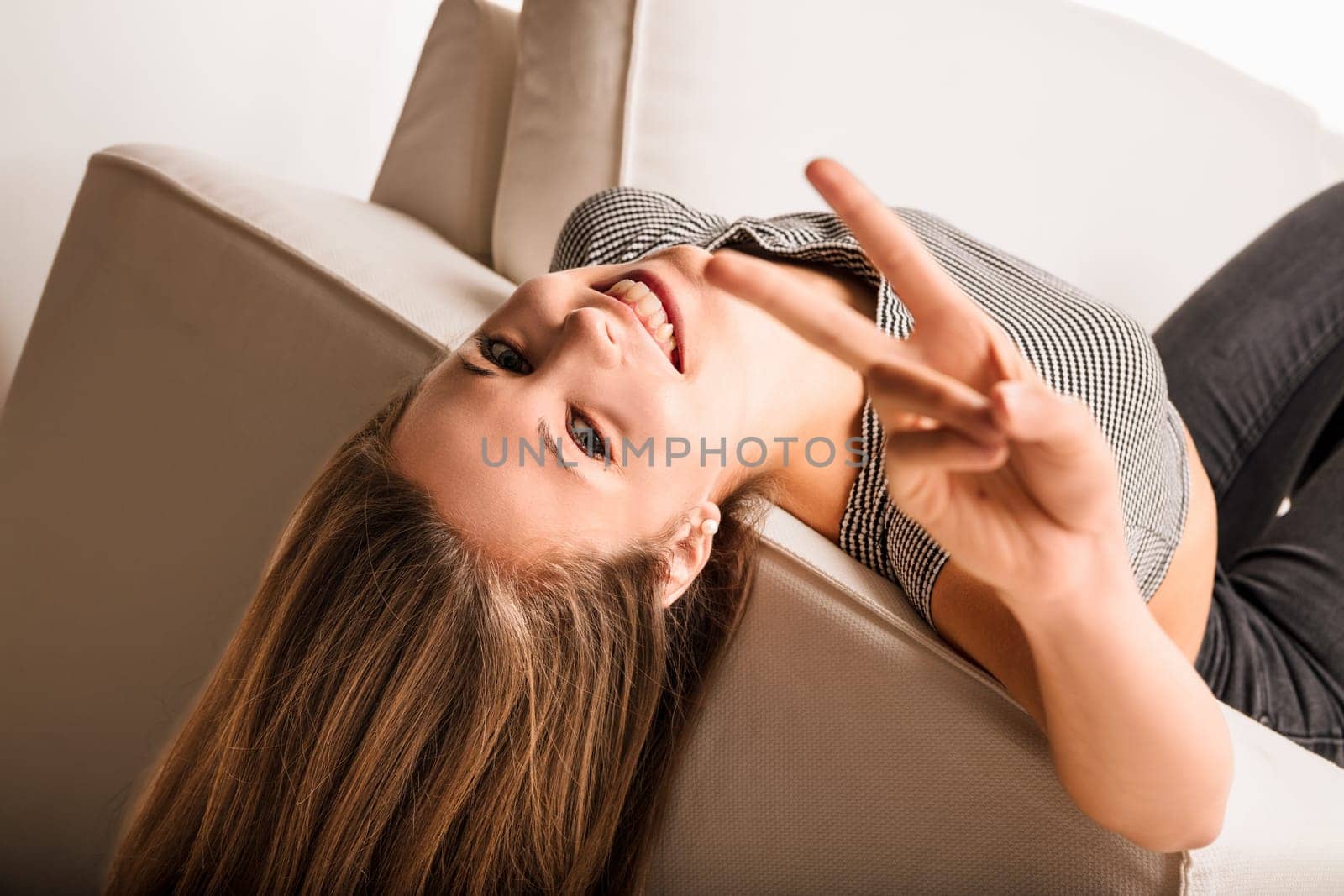 Teenage girl is lying upside down on a couch, smiling and gesturing a peace sign. Her long hair drapes over the back of the sofa