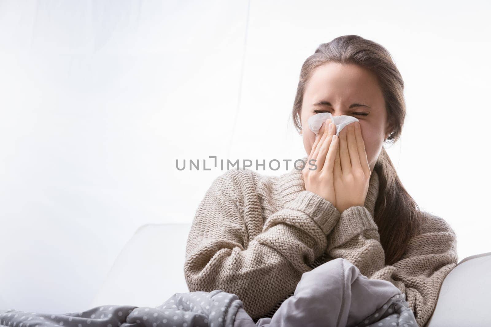 Sick young woman is sitting on her sofa, covered with a blanket, blowing her nose in a tissue. She is feeling unwell and has a headache