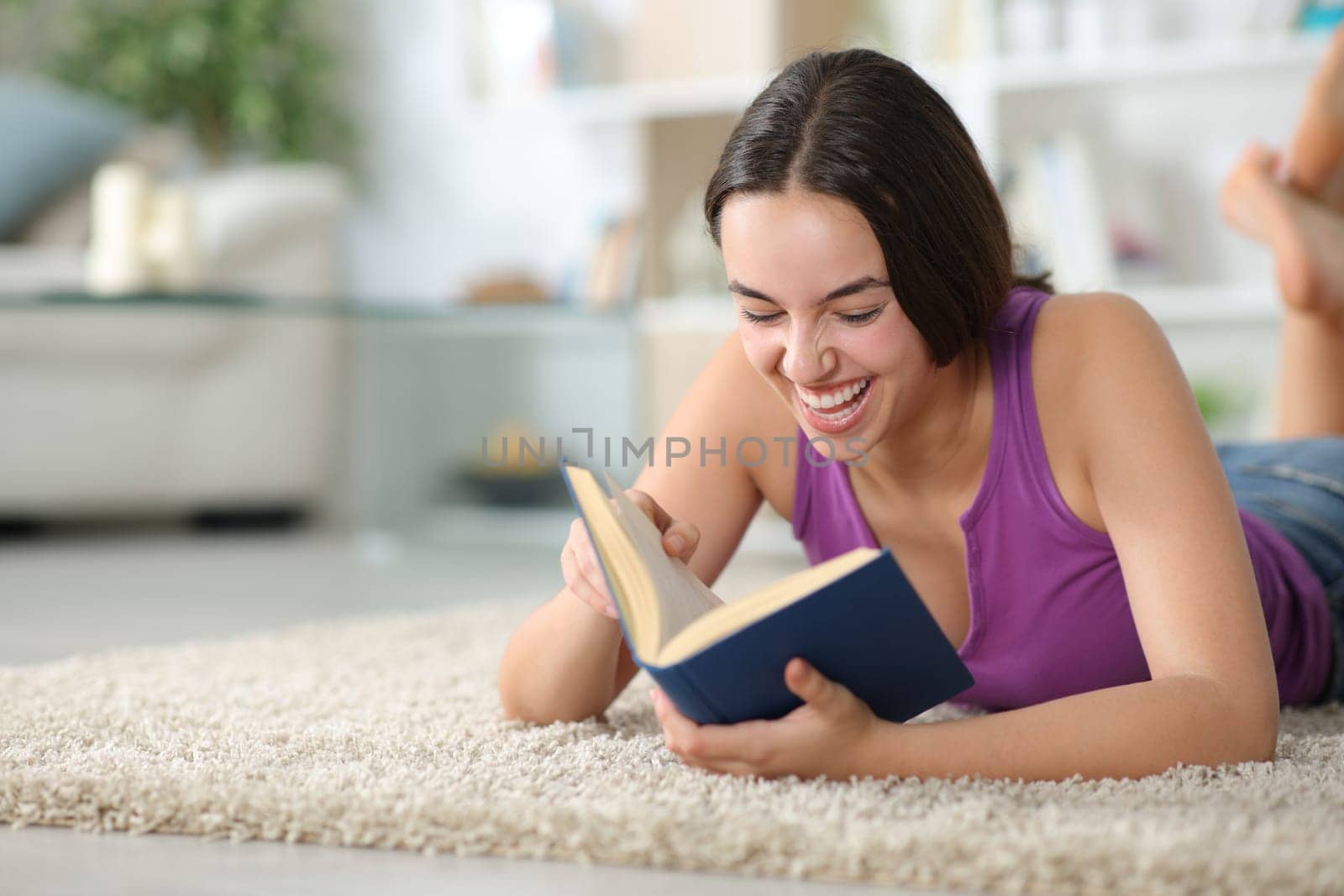 Happy woman laughing reading a paper book lying on a carpet at home