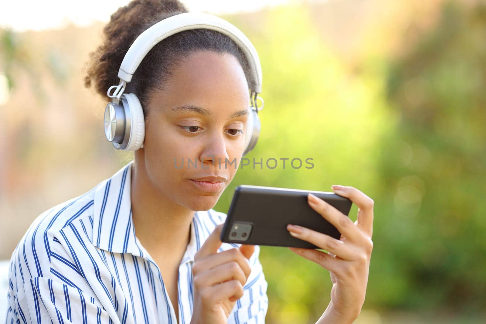 Serious black woman wearing headphone watching videos on horizontal cell phone in a park